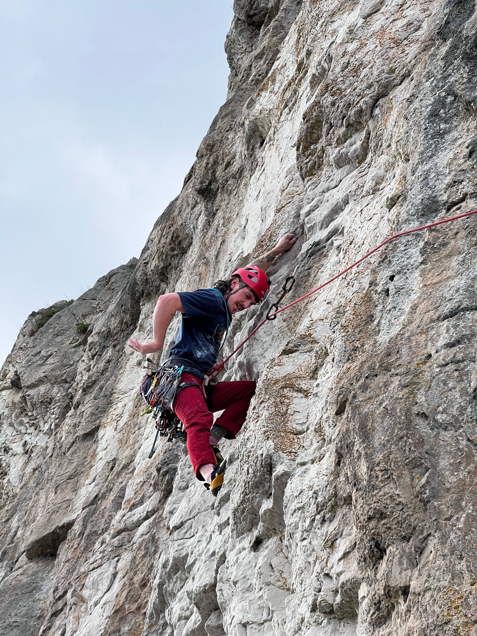 A person climbing a steep rock face, wearing a red helmet, climbing gear, and red pants, with a gray sky in the background.