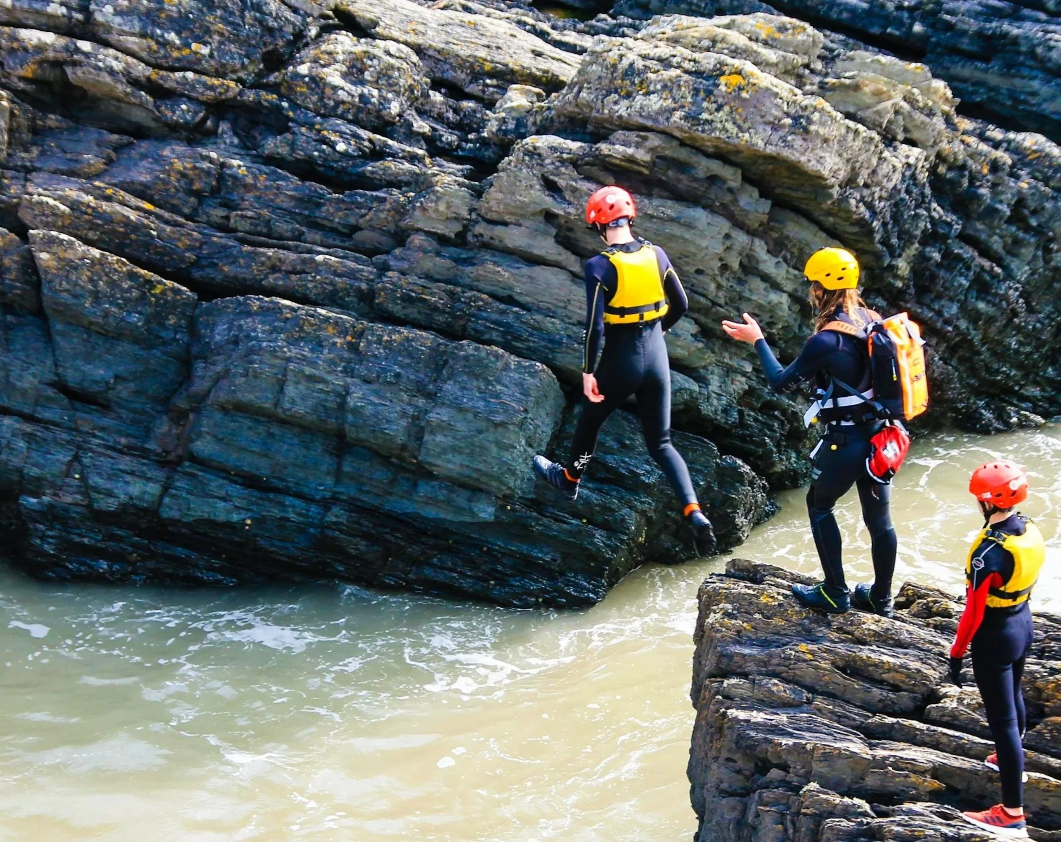 Three people wearing helmets and wetsuits crossing rocky terrain near water as part of a water adventure, with one individual helping another.