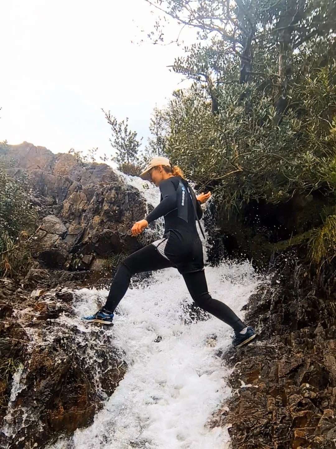 Person hiking or exploring a rocky stream with water cascading over rocks surrounded by vegetation.