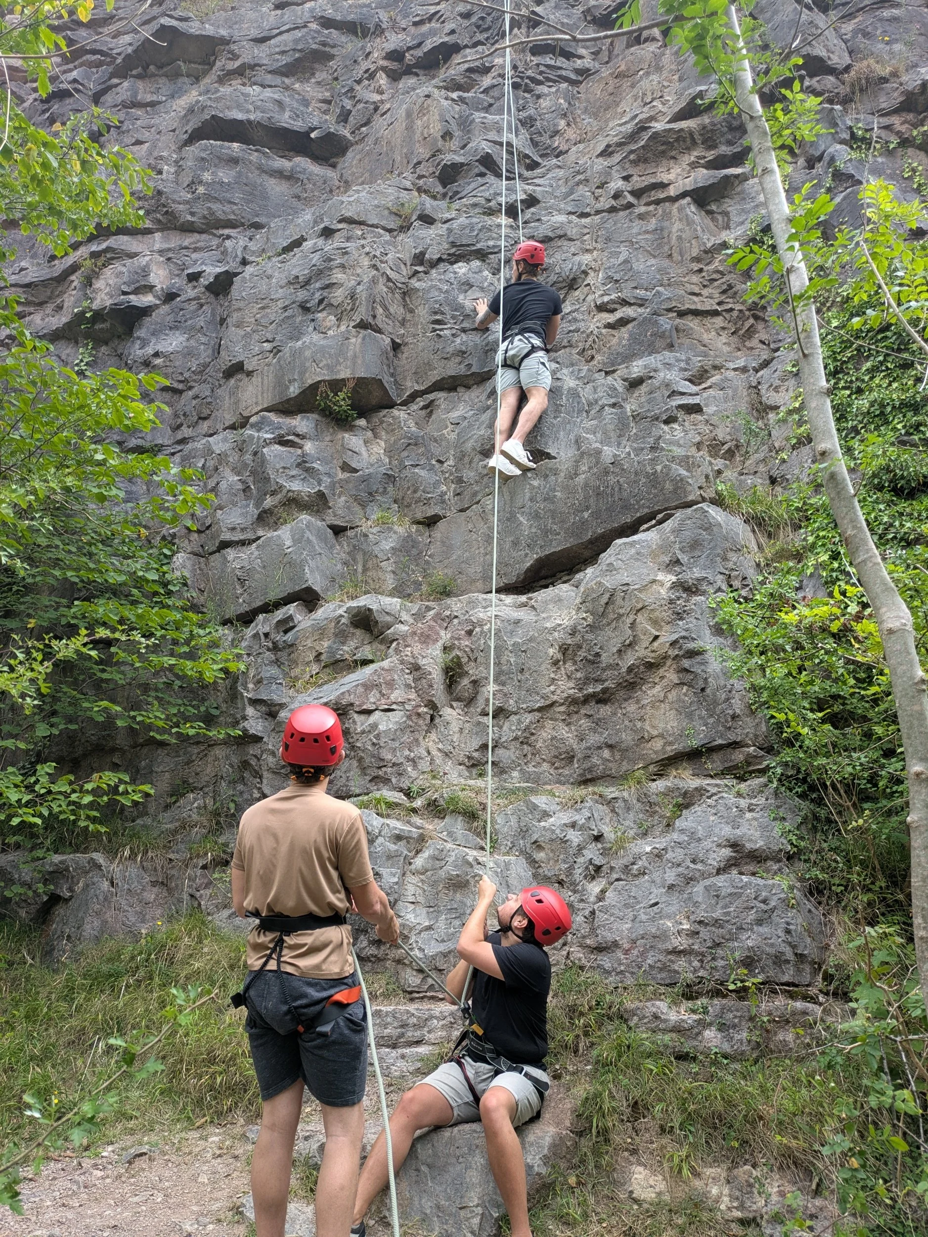 A person climbing a rock wall outdoors wearing a red helmet. Two individuals are standing below, assisting and supporting. The scene is surrounded by trees and greenery.