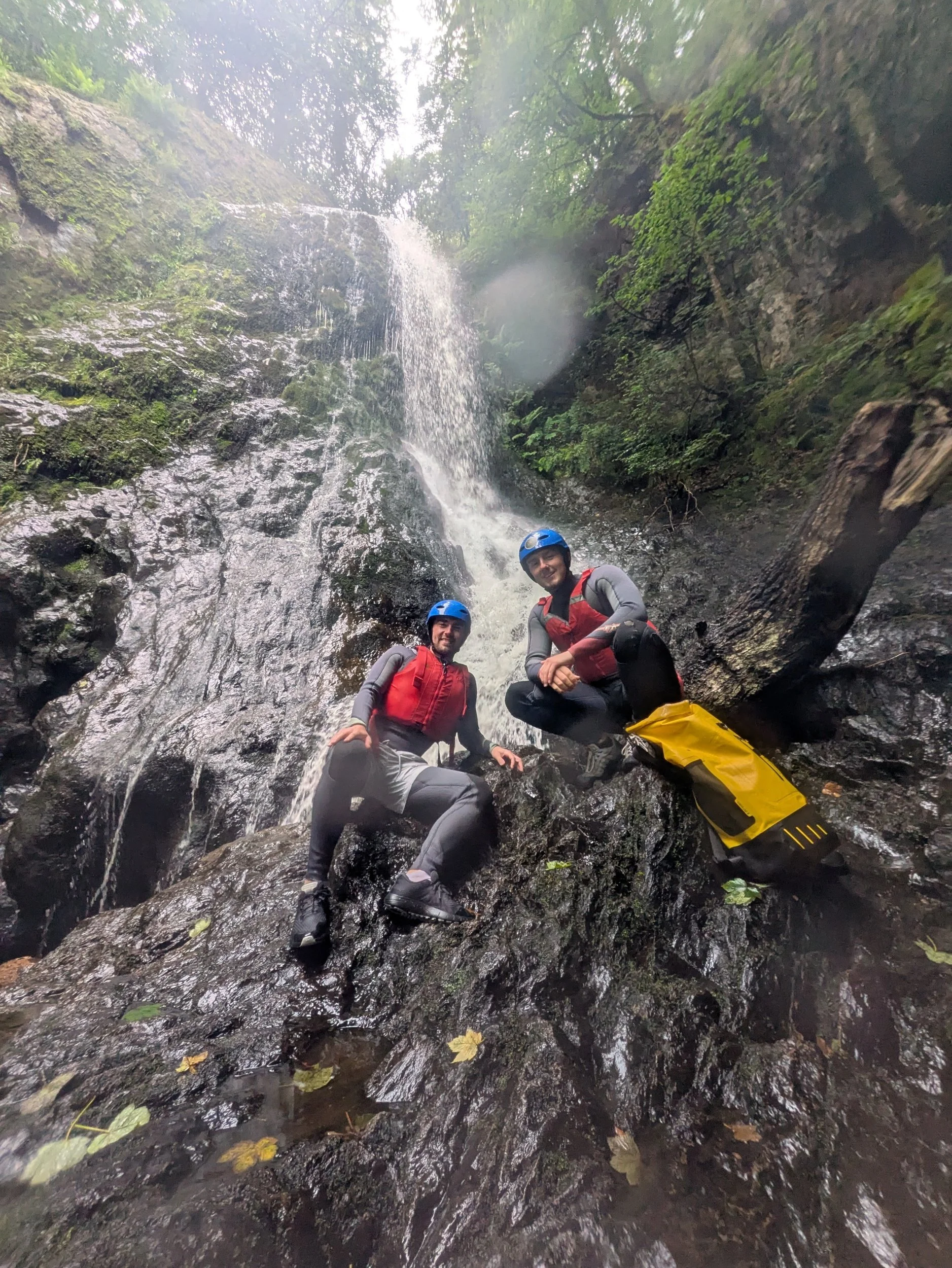Two people wearing helmets and waterproof gear near a waterfall in a lush green forest.