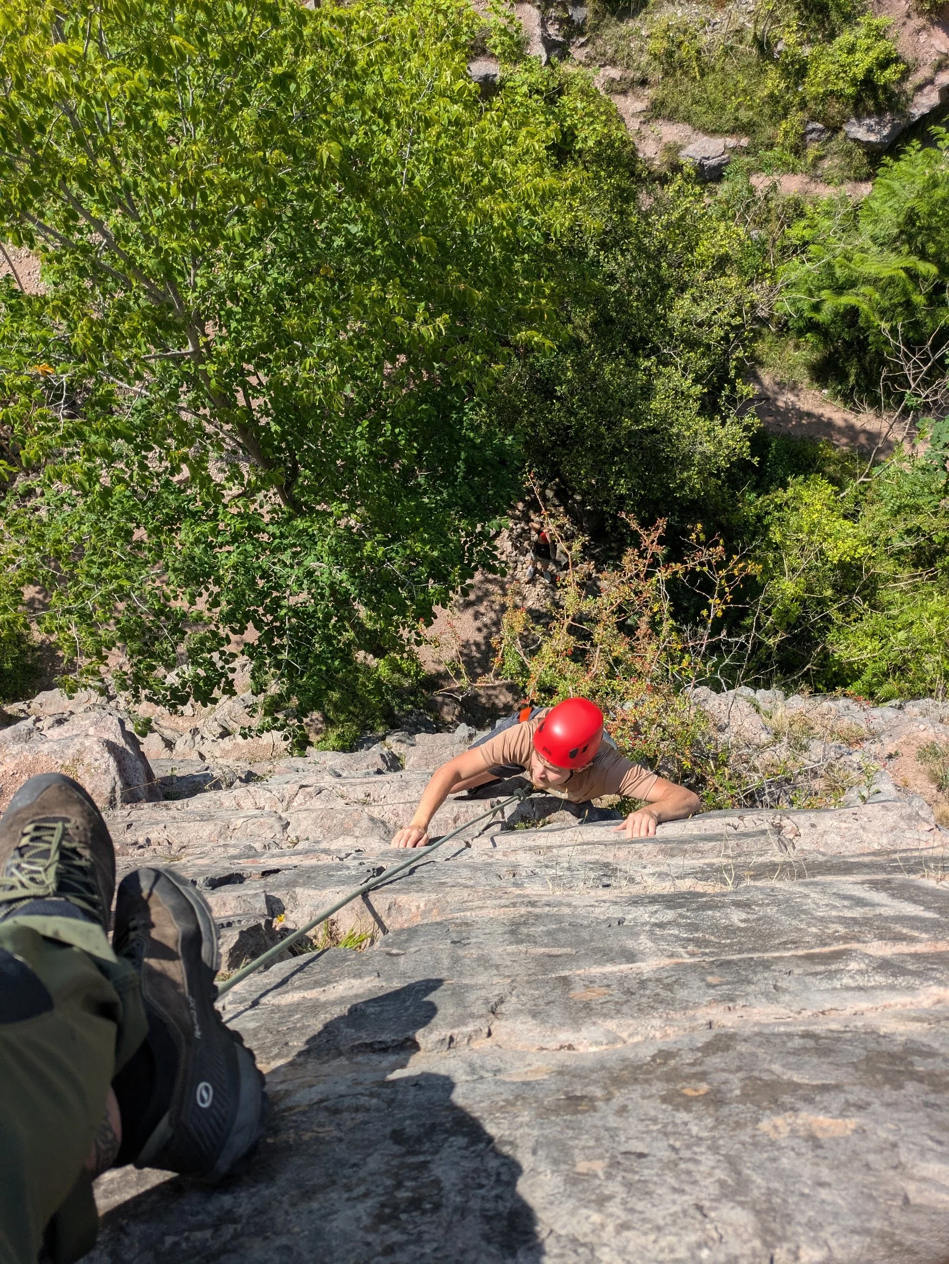 A person wearing a red helmet rock climbing on a steep rocky wall, viewed from above with the climber looking up and holding on to the rocks, surrounded by green trees and foliage.