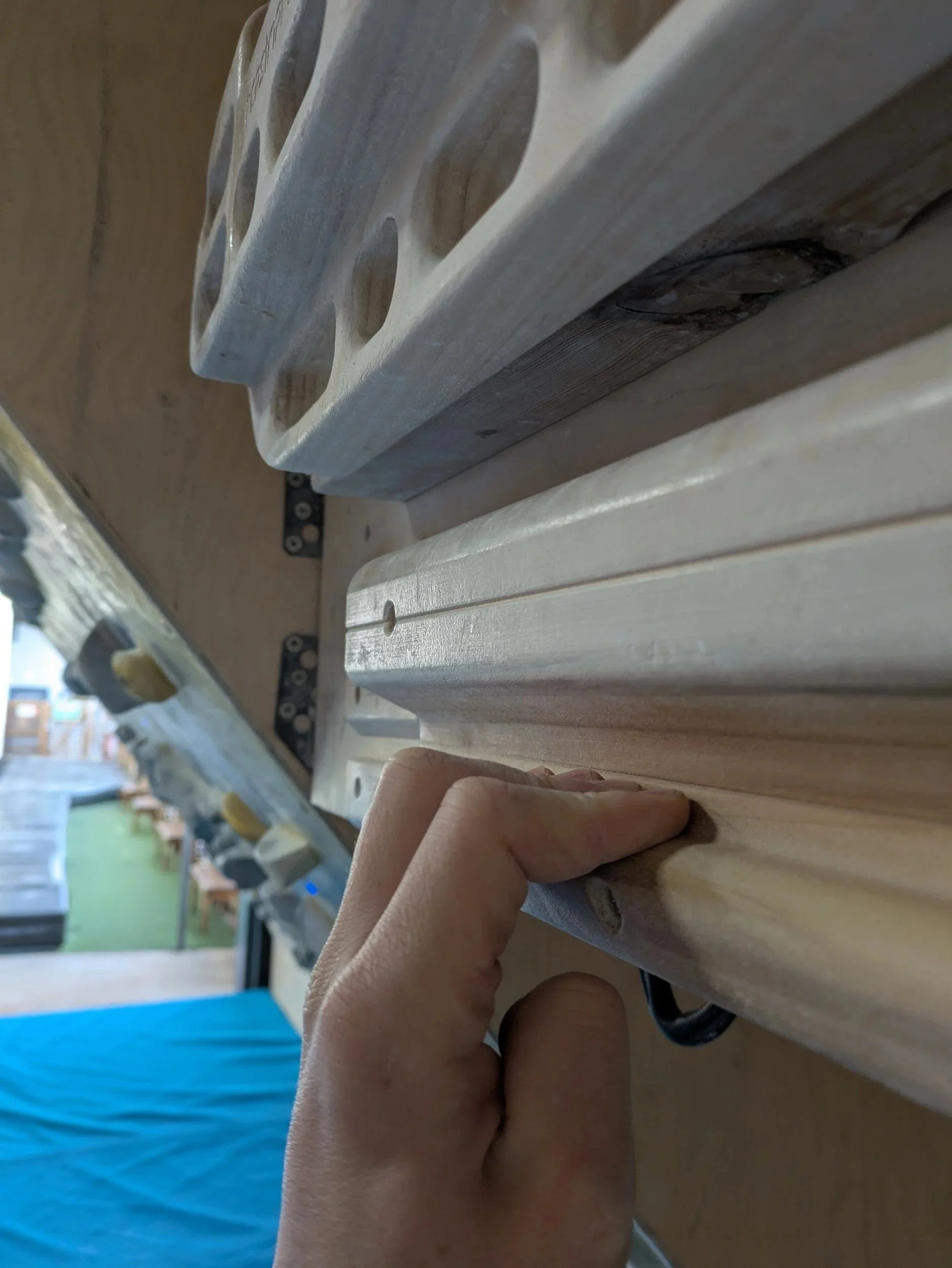 Close-up of a person's hand holding a strip of light-colored wood with a smooth, rounded edge, above a workbench in a workshop.