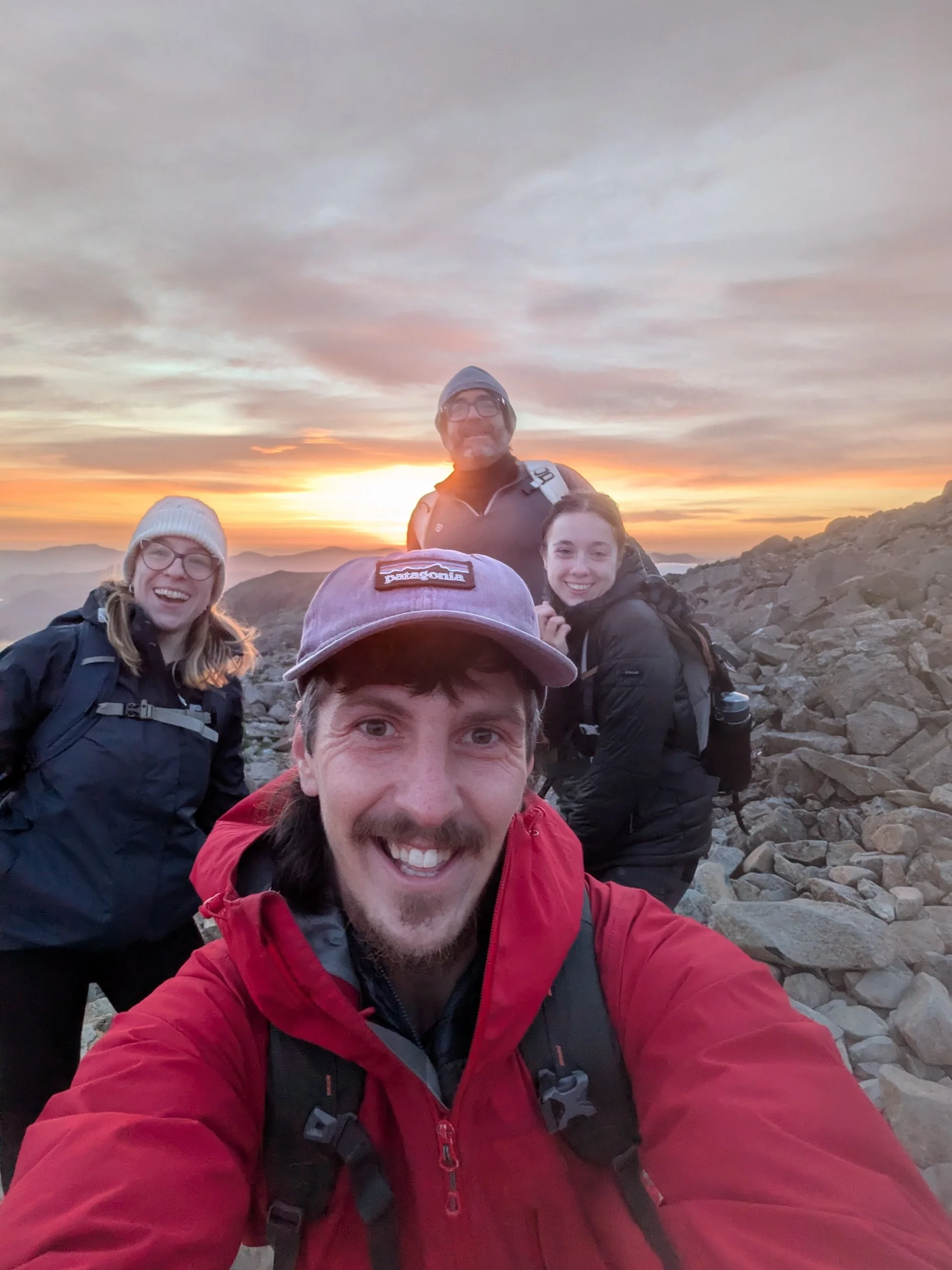 Group of four smiling hikers on rocky mountain summit at sunset, wearing outdoor gear and backpacks.