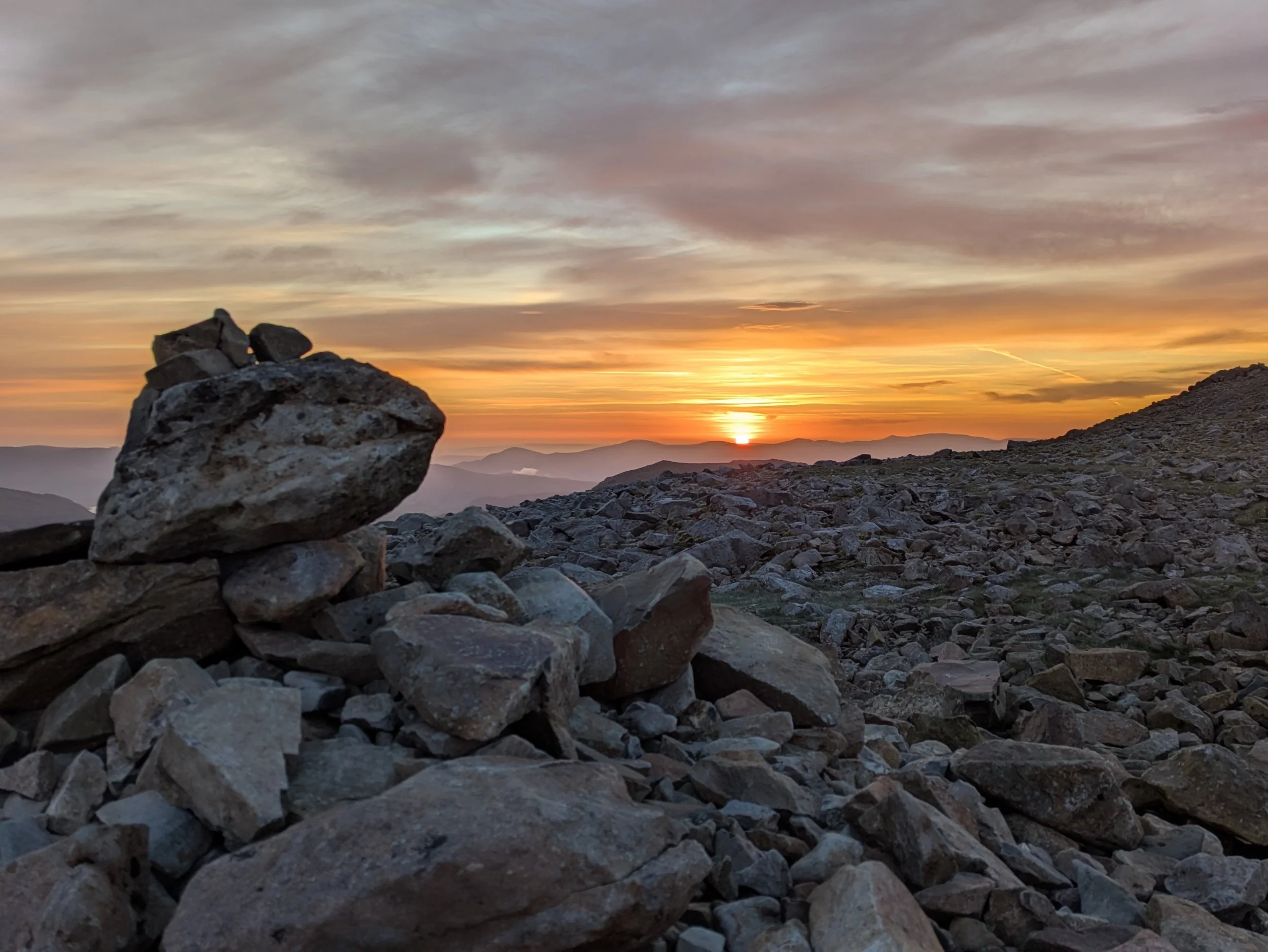 A pile of rocks and stones in the foreground at sunset with a colorful sky, clouds, and mountains in the distance.
