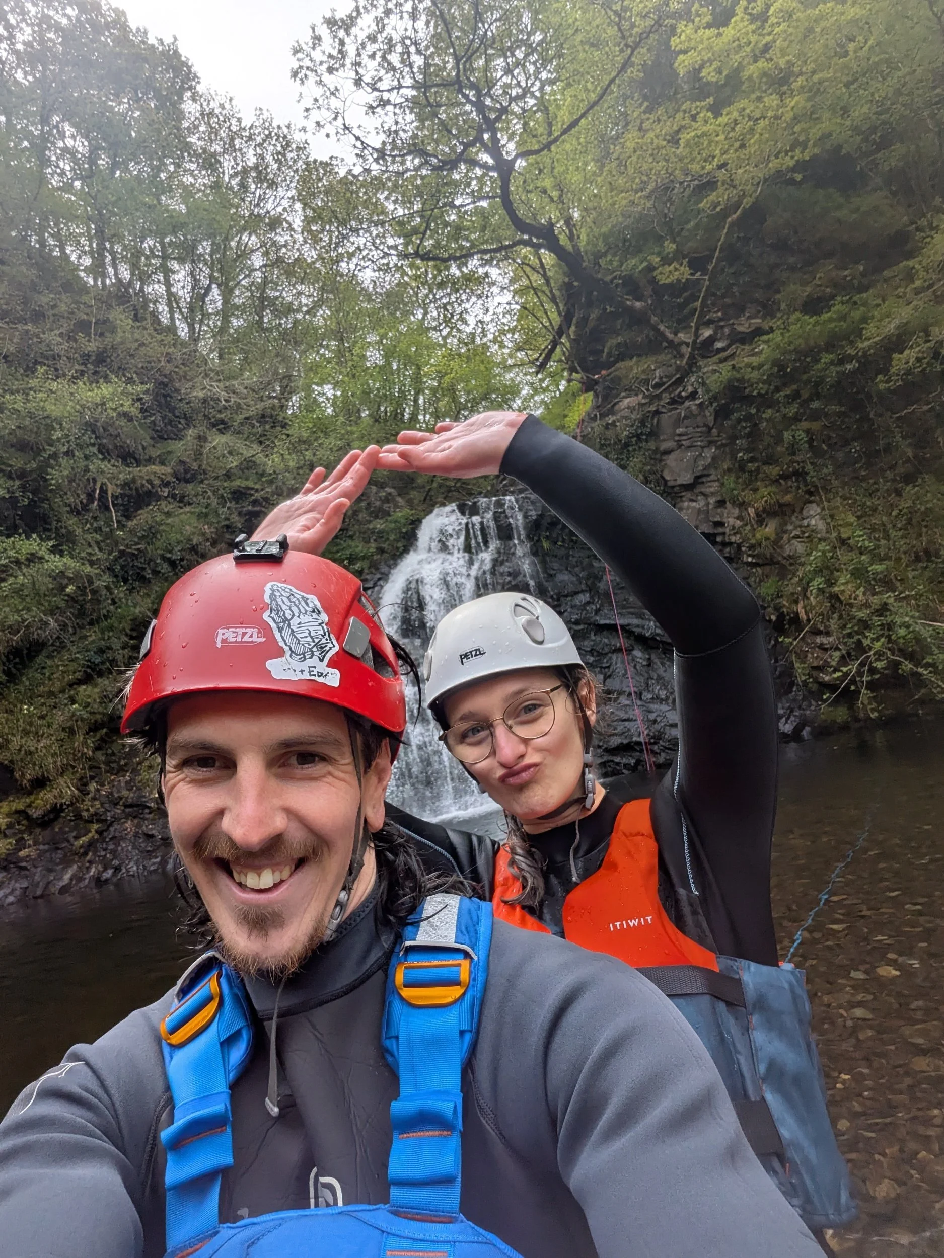 Two people in helmets and outdoor gear taking a selfie in front of a waterfall in a forest.