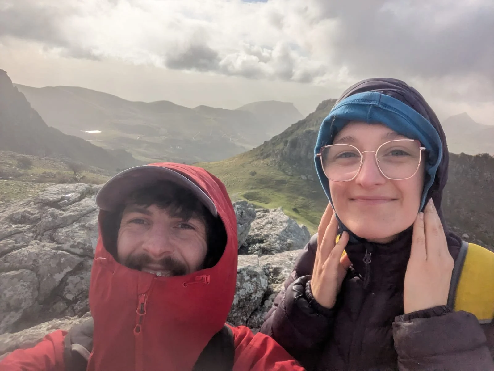 Two hikers wearing jackets and head coverings taking a selfie on a mountain with rocky terrain, lush green valleys, and distant mountains under cloudy skies in the background.