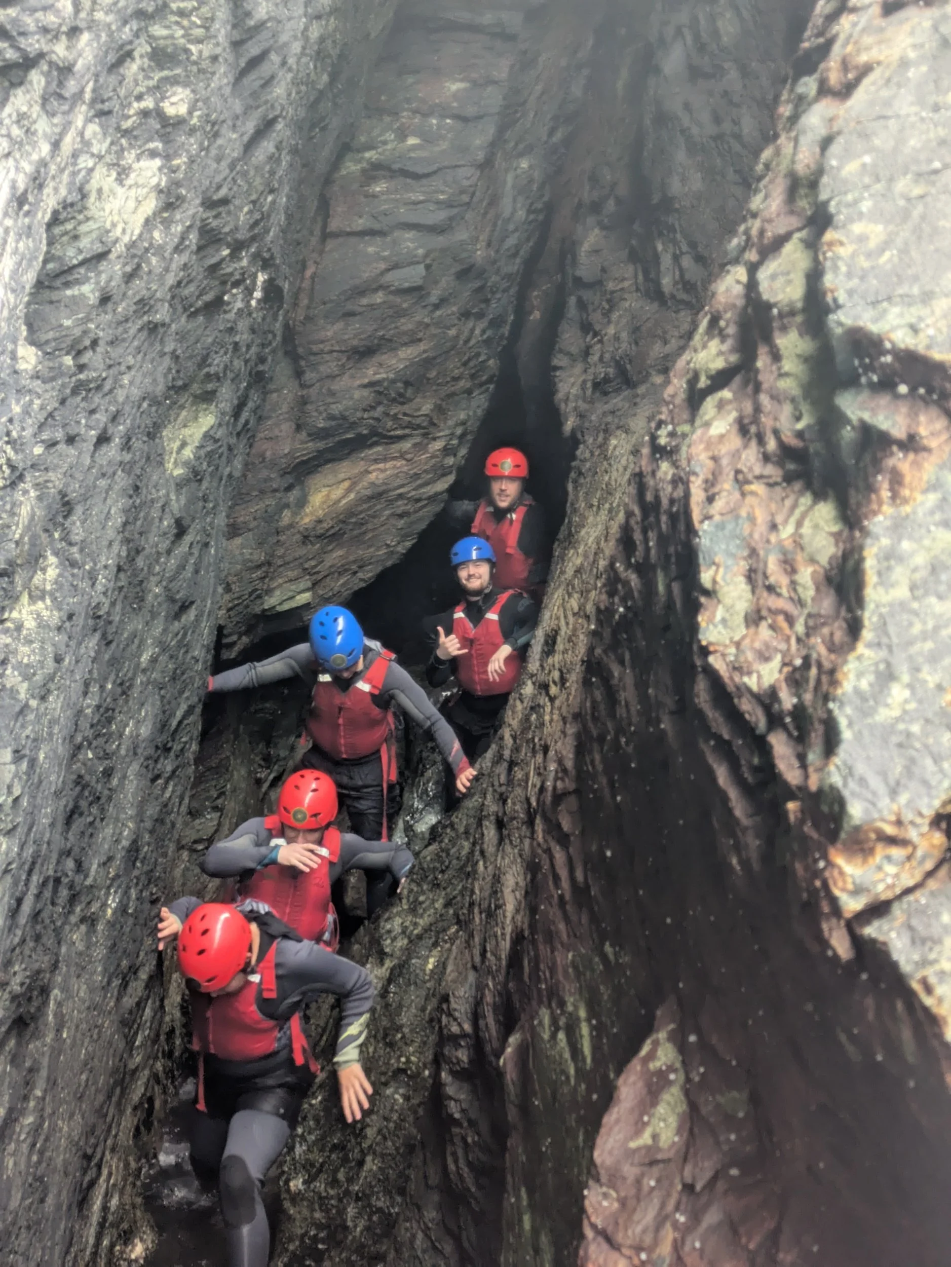 A group of people climbing a narrow, rocky crevice outdoors, wearing helmets and safety gear.