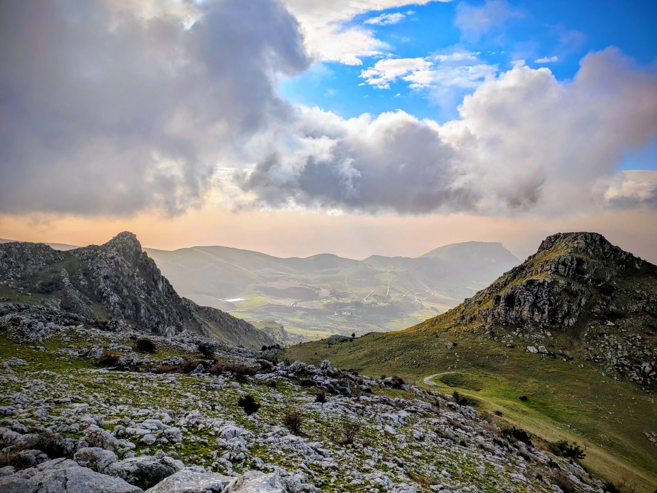 Mountain landscape with rocky foreground, rolling hills in the distance, and a partly cloudy sky with patches of blue.