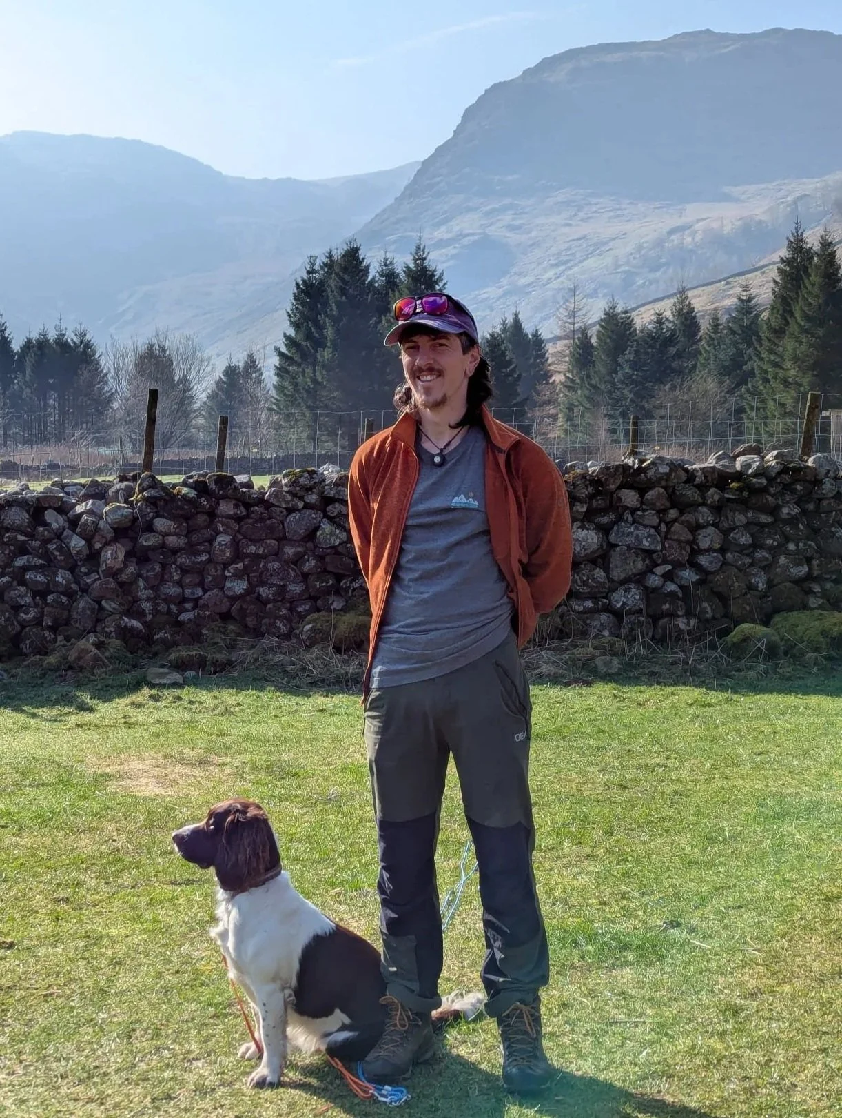 A man with dark brown hair, mustache, and goatee, wearing a navy cap with sunglasses on top, a brown jacket, gray t-shirt, dark pants, and hiking boots, standing on a grassy area with a dog sitting beside him. The dog is a spaniel with black and white fur, and a leash attached to its collar. Behind them is a stone wall with trees and mountains in the background, and the weather appears clear and sunny.