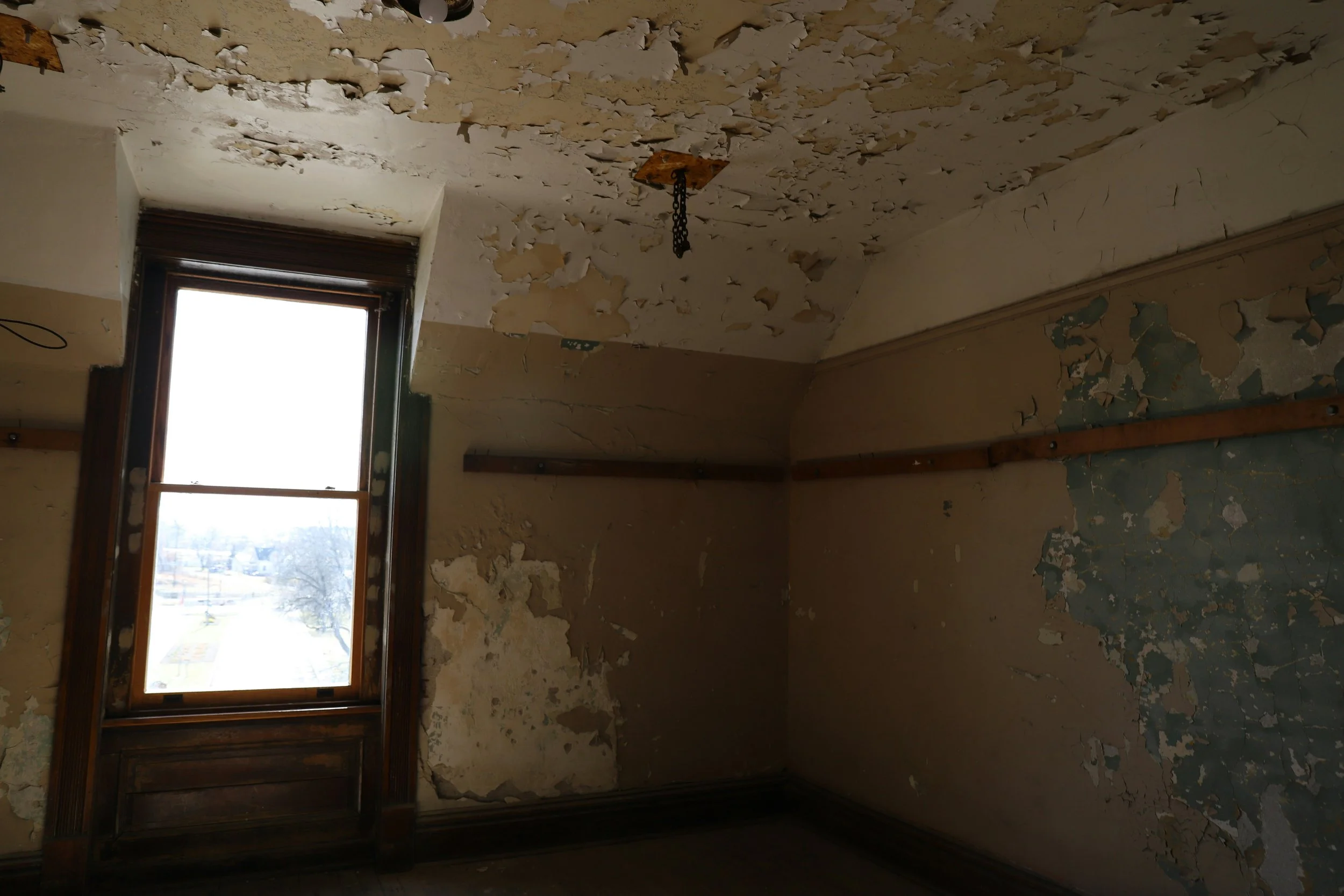 A room with peeling paint and damaged walls, featuring an open window letting in natural light, with exposed wood trim and a light fixture hanging from the ceiling.