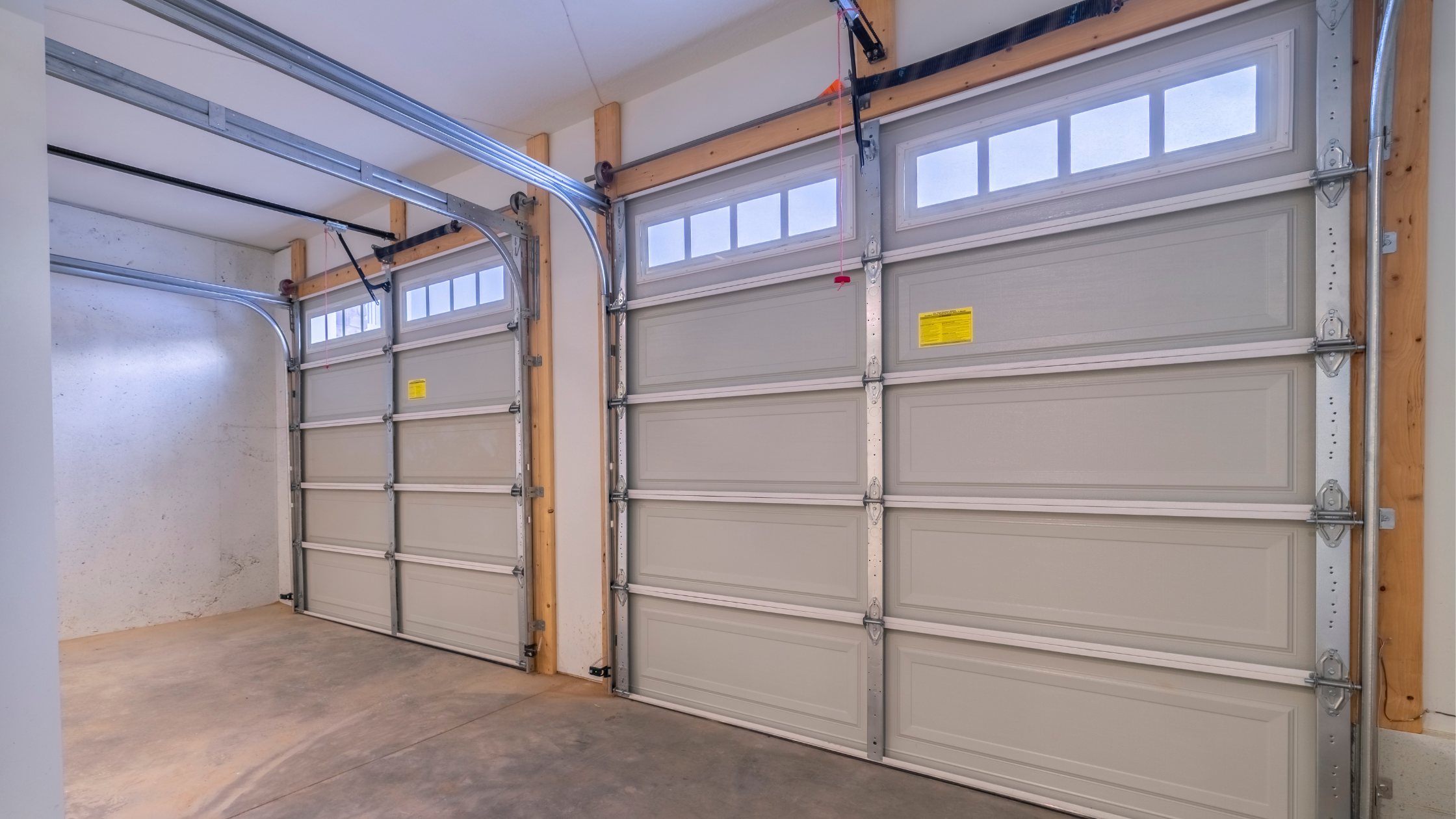 Two paneled garage doors with metal tracks, rollers, and a partial view of glass windows at the top, inside a garage with unfinished walls and concrete floor.