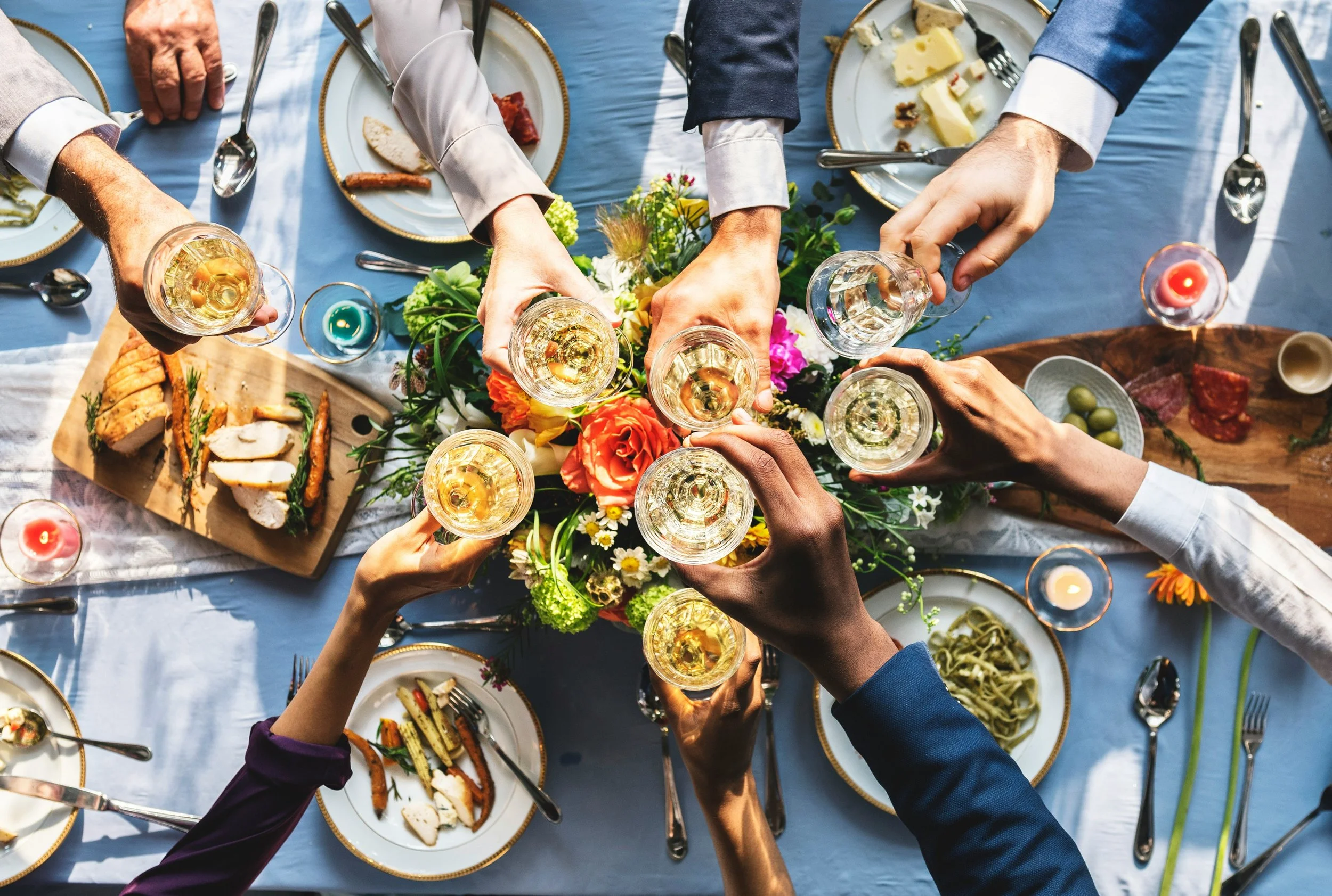 Persone che brindano con bicchieri di vino bianco in un tavolo apparecchiato con cibo e un centrotavola di fiori colorati.