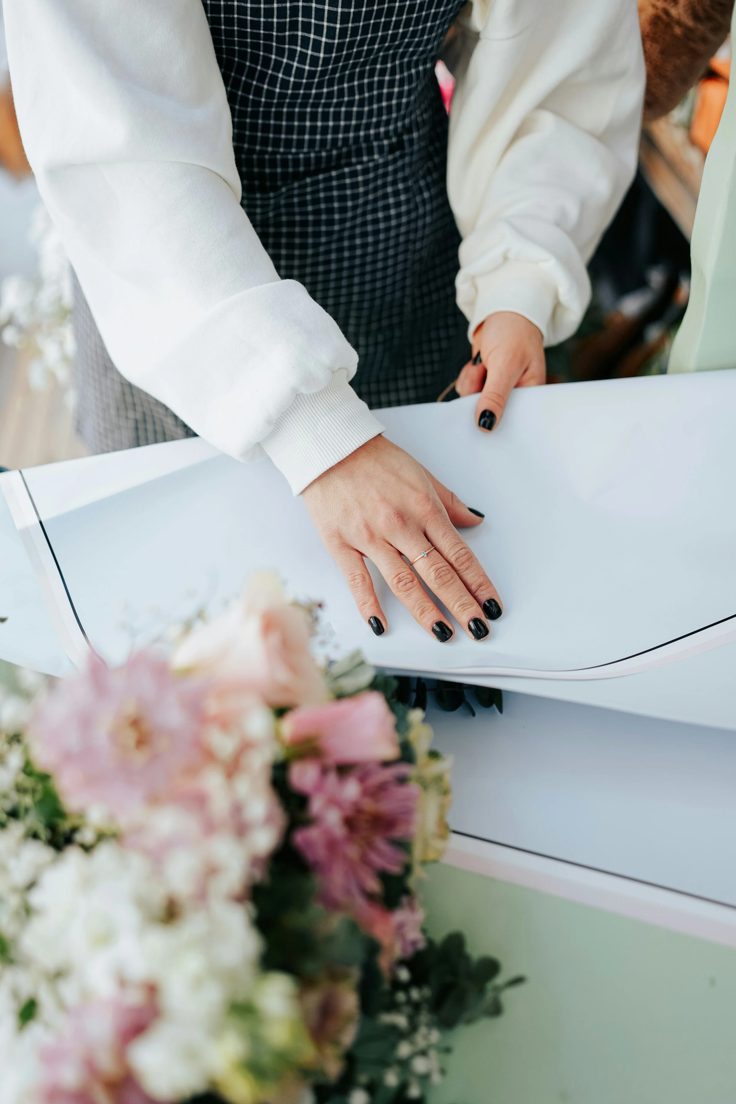 Una persona con un anello sulla mano sta firmando un documento, con un mazzo di fiori sfocato in primo piano.