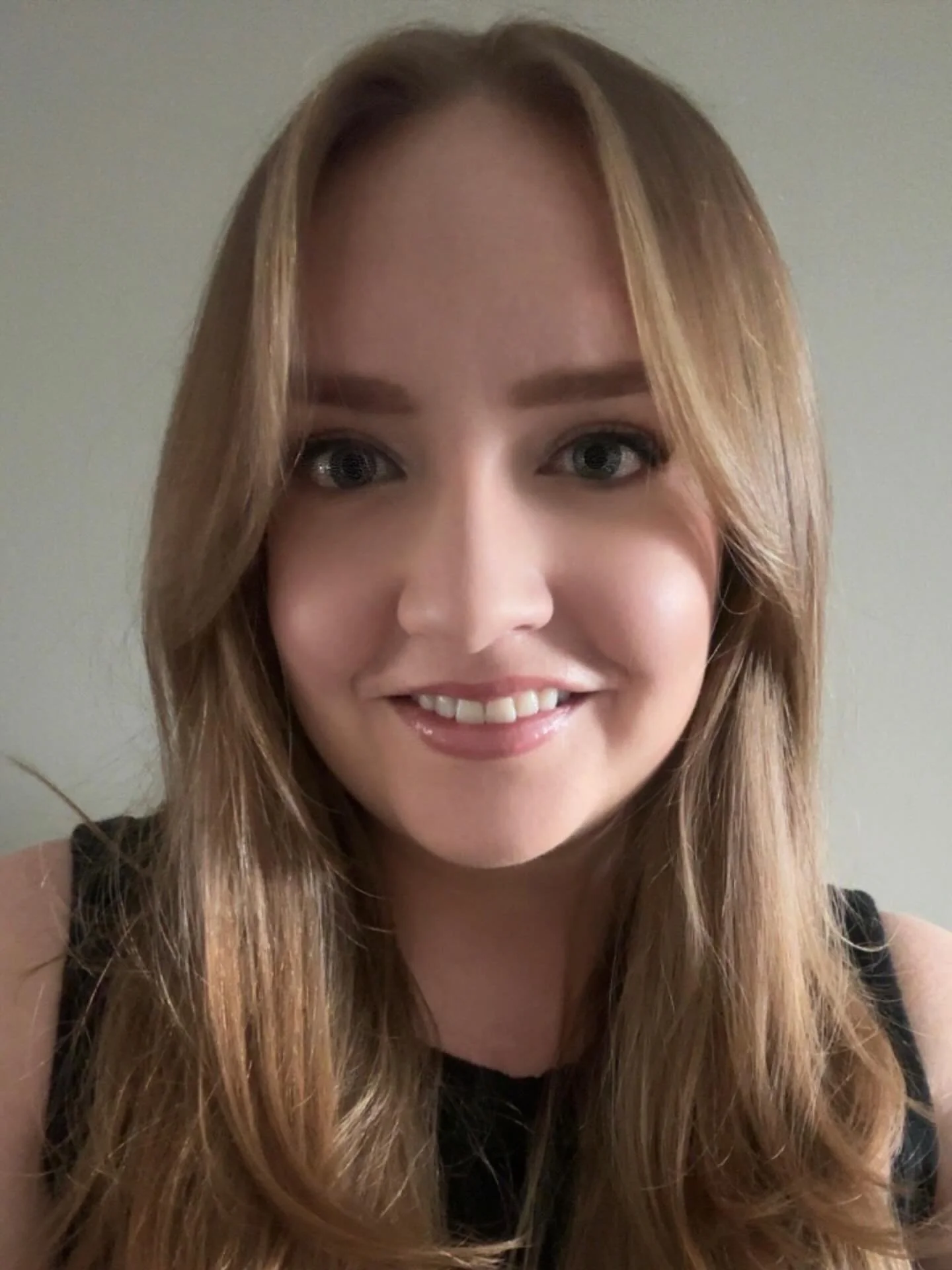 A woman with long, wavy blonde hair smiling at the camera, wearing a black top, in front of a plain wall.