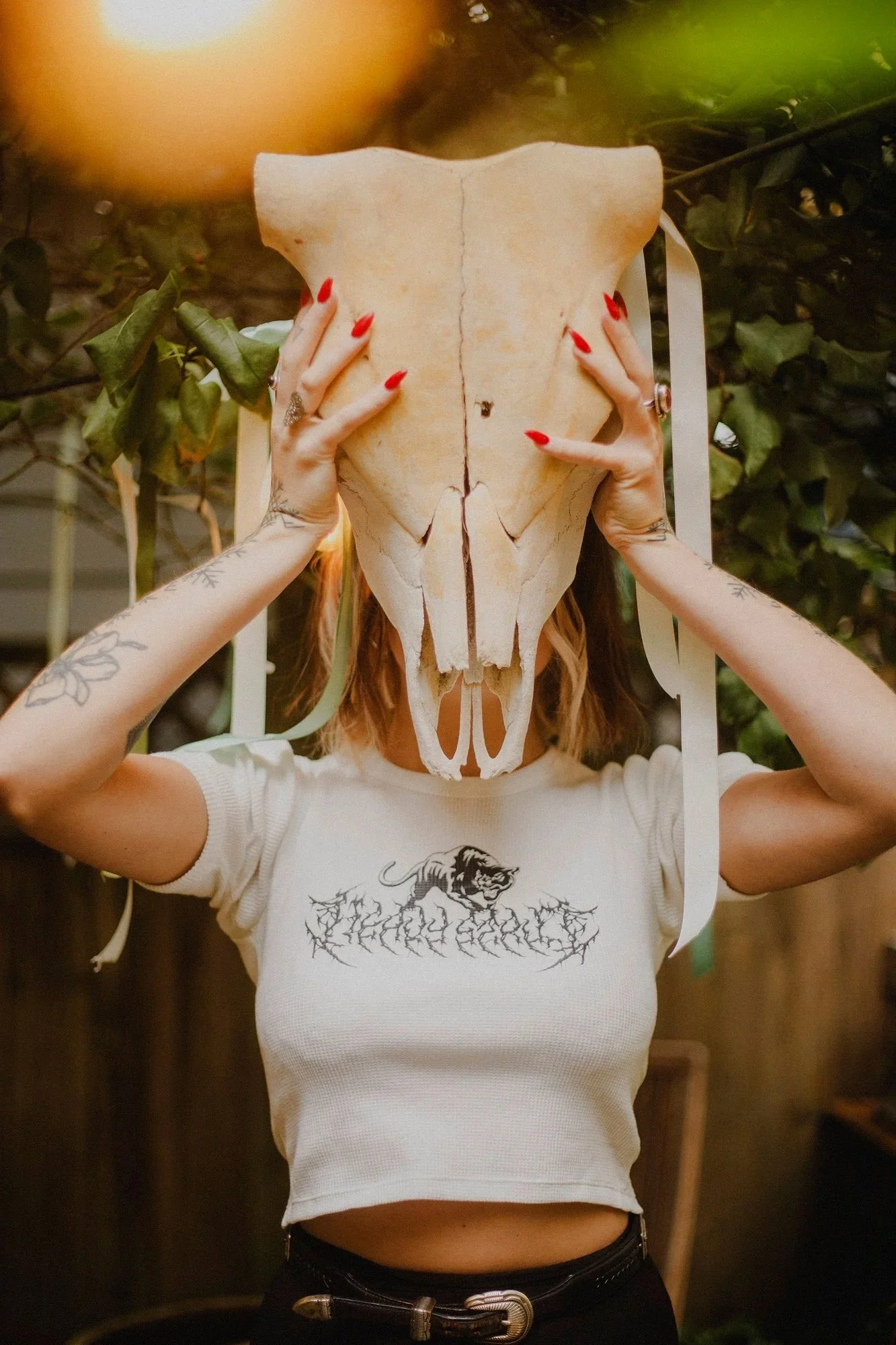Person holding a large animal skull mask in front of their face, with tattoos visible on their arms. The person is wearing a white t-shirt with a lion graphic and black pants with a belt. The background has greenery.