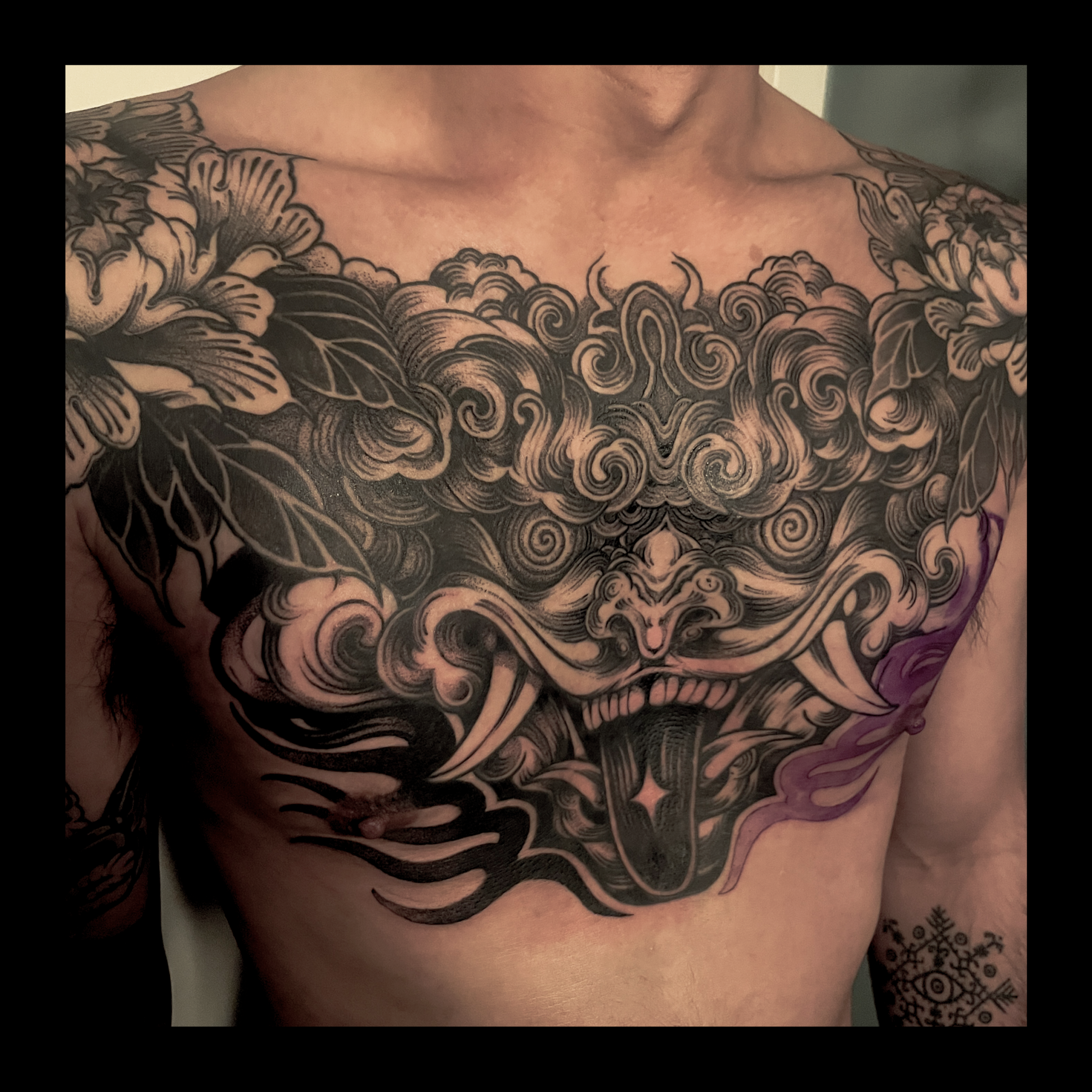 Close-up of a person's upper chest and shoulders with intricate black and gray tattoos featuring floral, swirl, and abstract patterns.