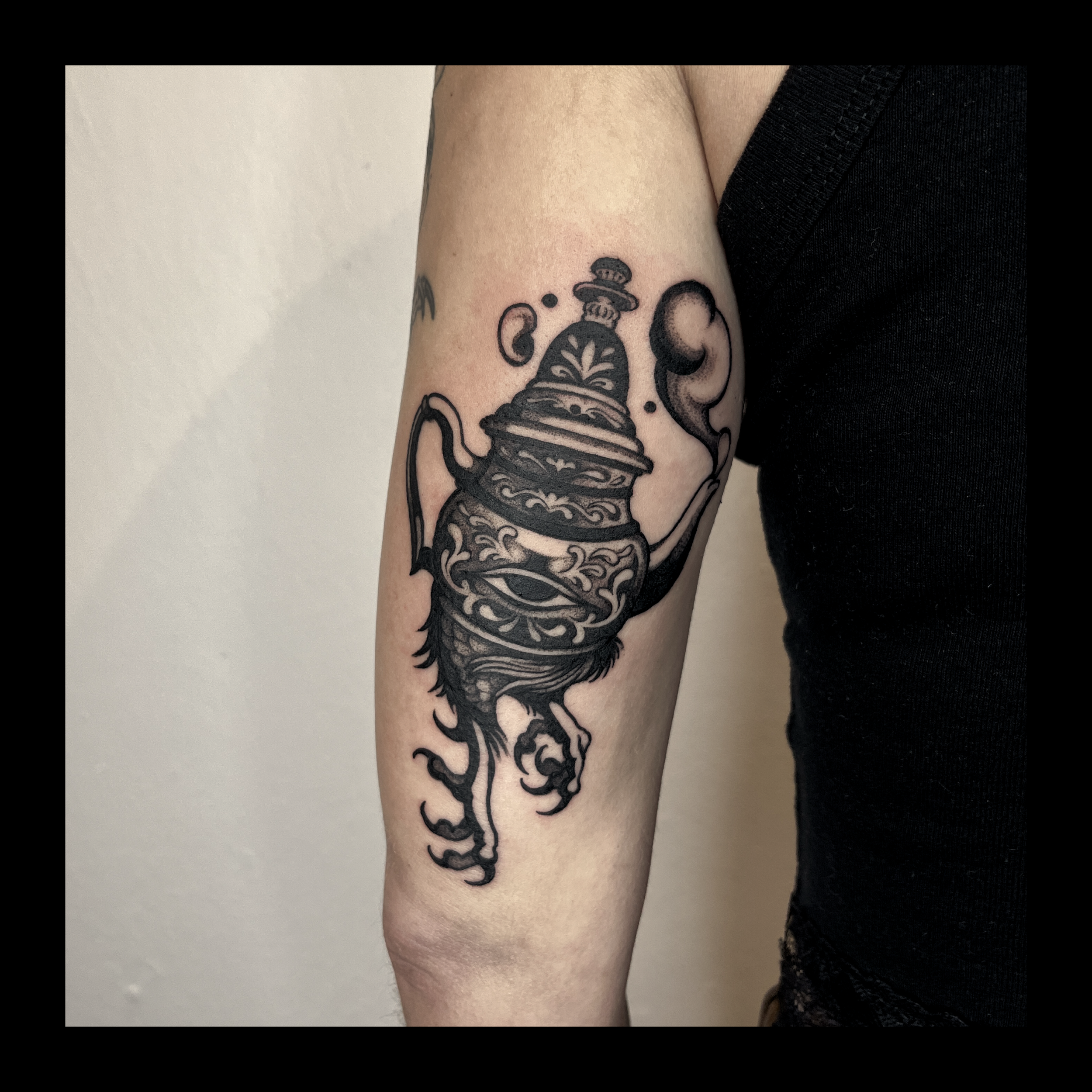 Close-up of a person's upper arm with a black tattoo of a decorative teapot and floral designs, wearing a black sleeveless top.