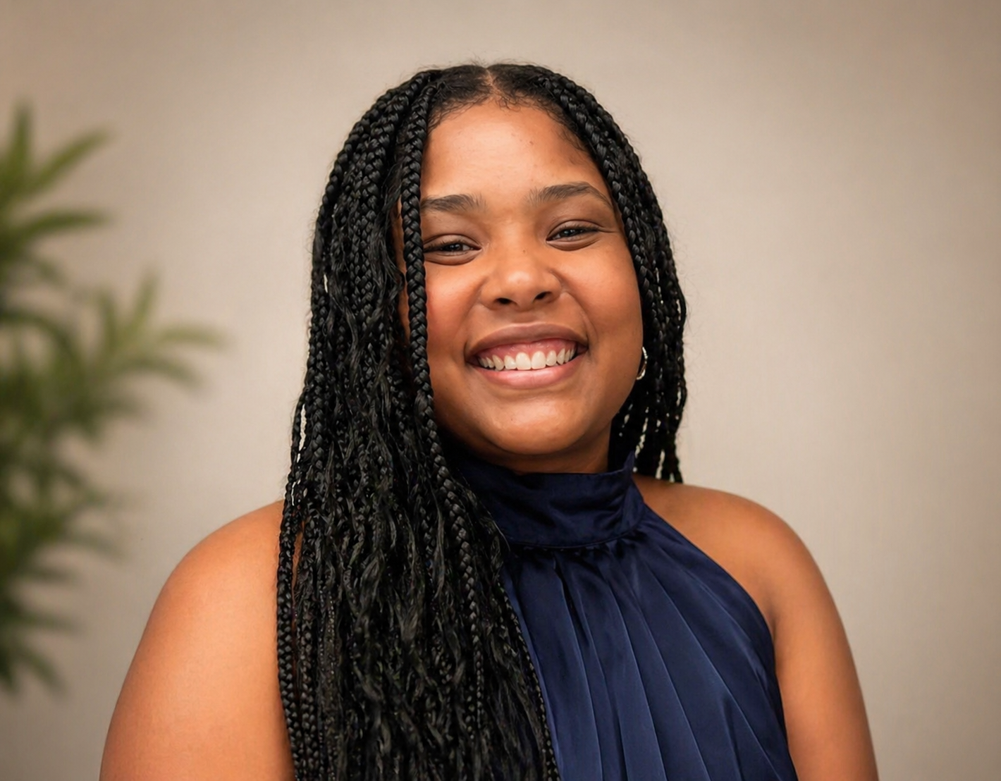 A smiling young woman with long black braids wearing a sleeveless dark blue top, standing against a plain background.