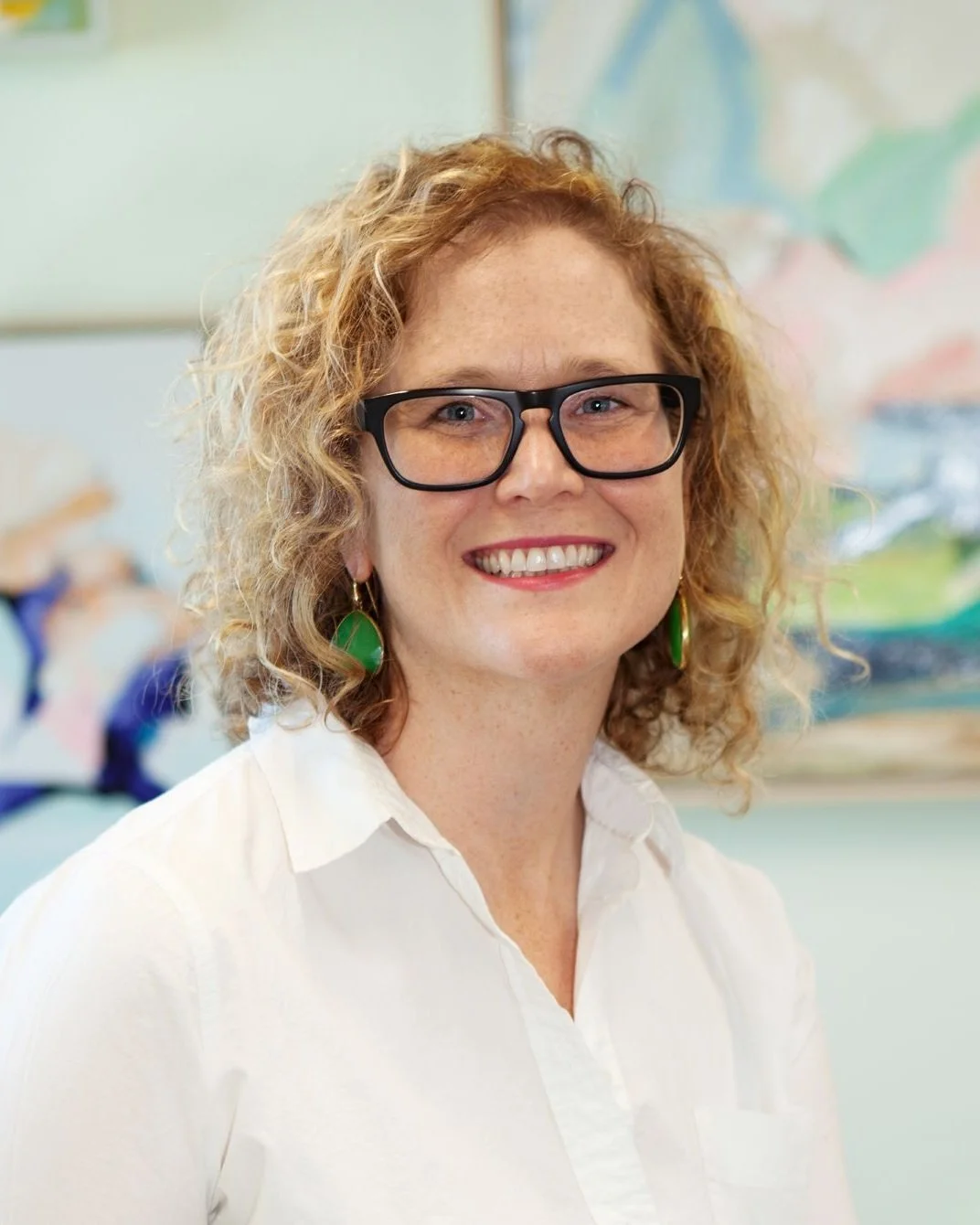 A woman with curly red hair, glasses, and green earrings, smiling in front of a colorful abstract background.