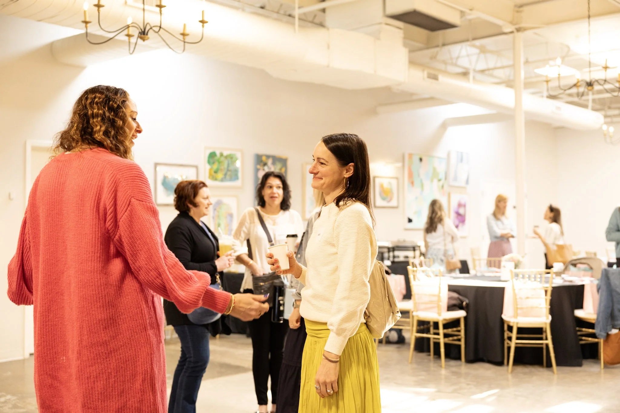 Two women are smiling and talking to each other at an indoor social event. One woman has curly hair and is wearing a pink sweater, and the other has straight dark hair and is wearing a light-colored top and yellow skirt. Other people are visible in t