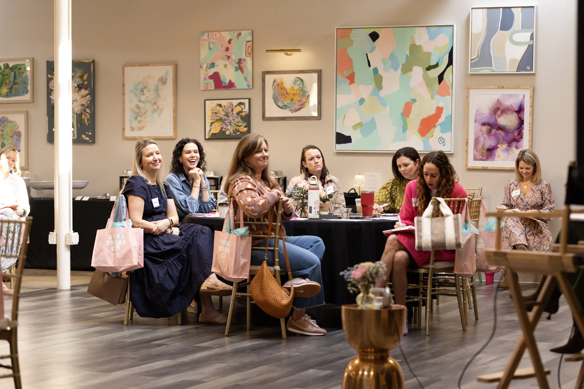Women attending an art event or workshop in a gallery space with abstract paintings on the wall, some women are smiling and others are focused on activities, with gift bags on the table.