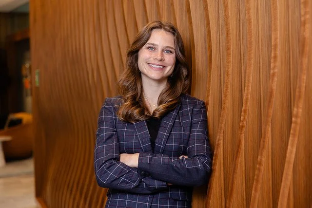 A woman with brown hair and a plaid blazer smiling and standing with her arms crossed against a wooden wall in an indoor setting.