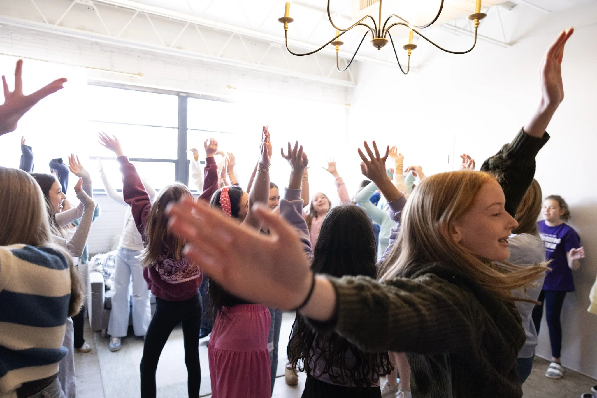 A group of children raising their hands enthusiastically indoors near a large window and a modern chandelier.
