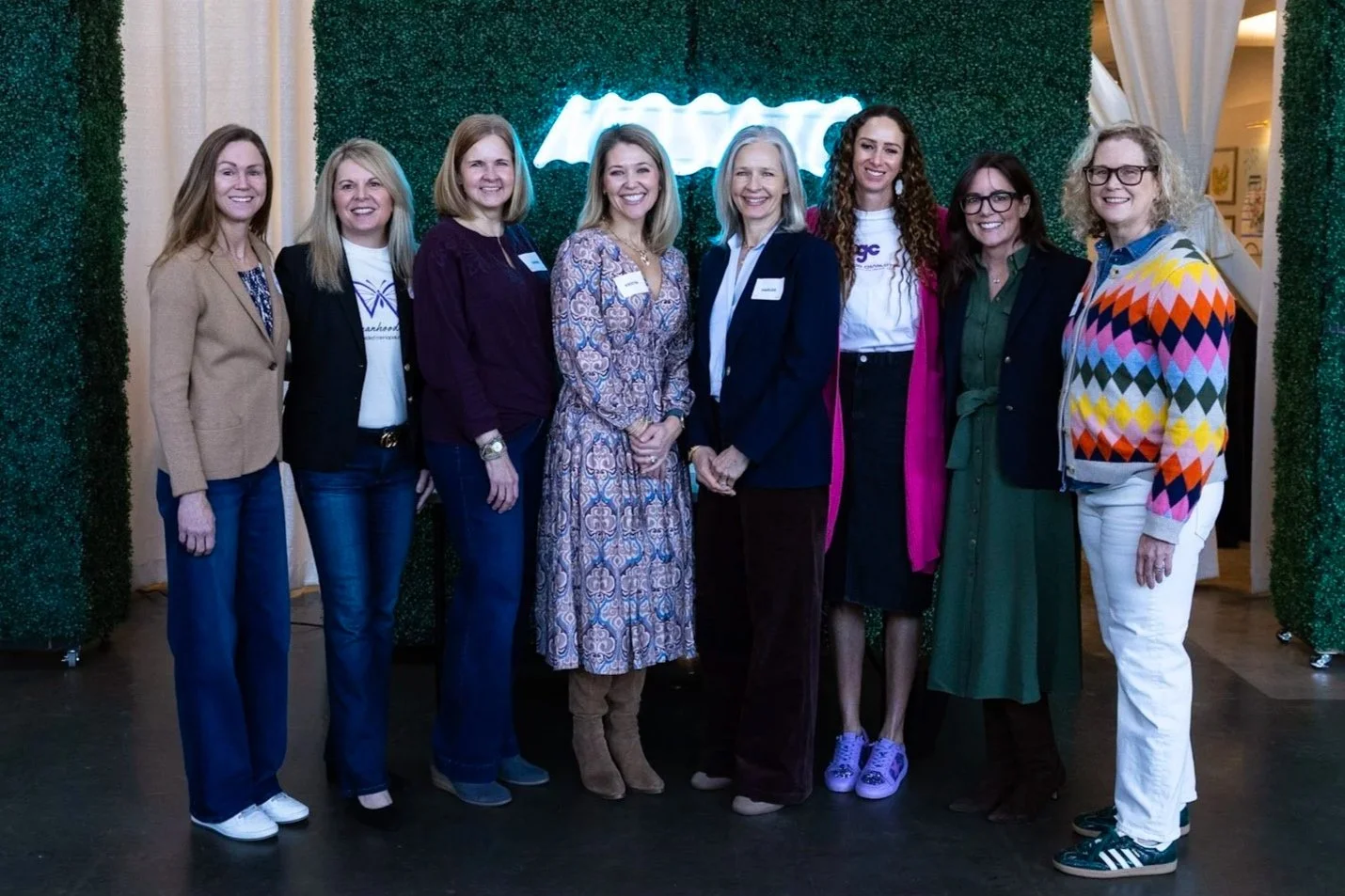 Group of nine women standing together in front of a green backdrop and neon sign at an indoor event.