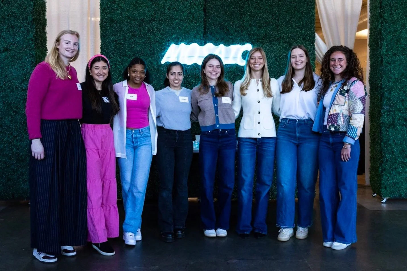 Group of nine young women standing together indoors in front of a green hedge wall with a neon sign. They are smiling and dressed casually in colorful and neutral outfits.