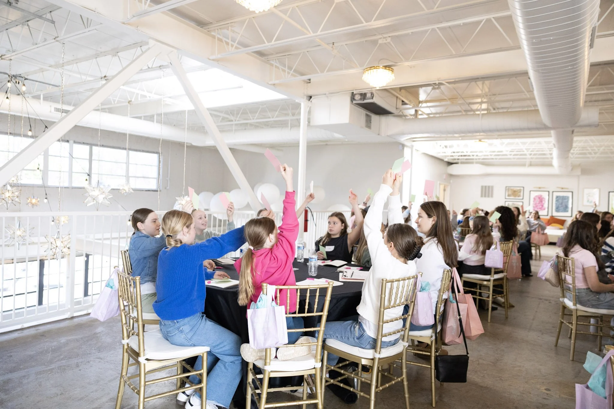 A group of young girls sitting around a table in a bright, industrial-style event space, raising their hands and holding colored paper in a classroom or workshop setting.