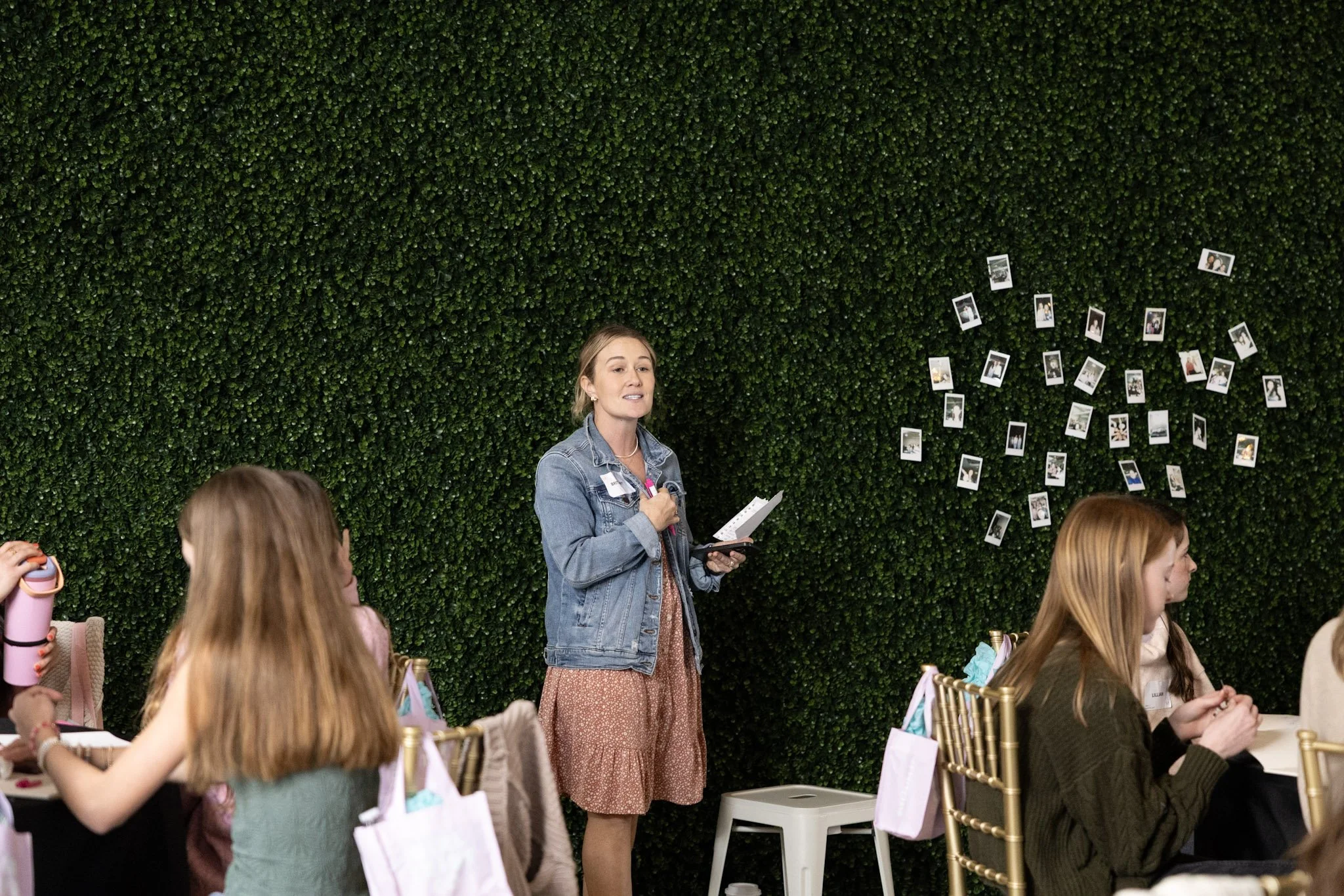 A woman in a denim jacket speaking to a group of children during an event, with a green wall displaying photos behind her.