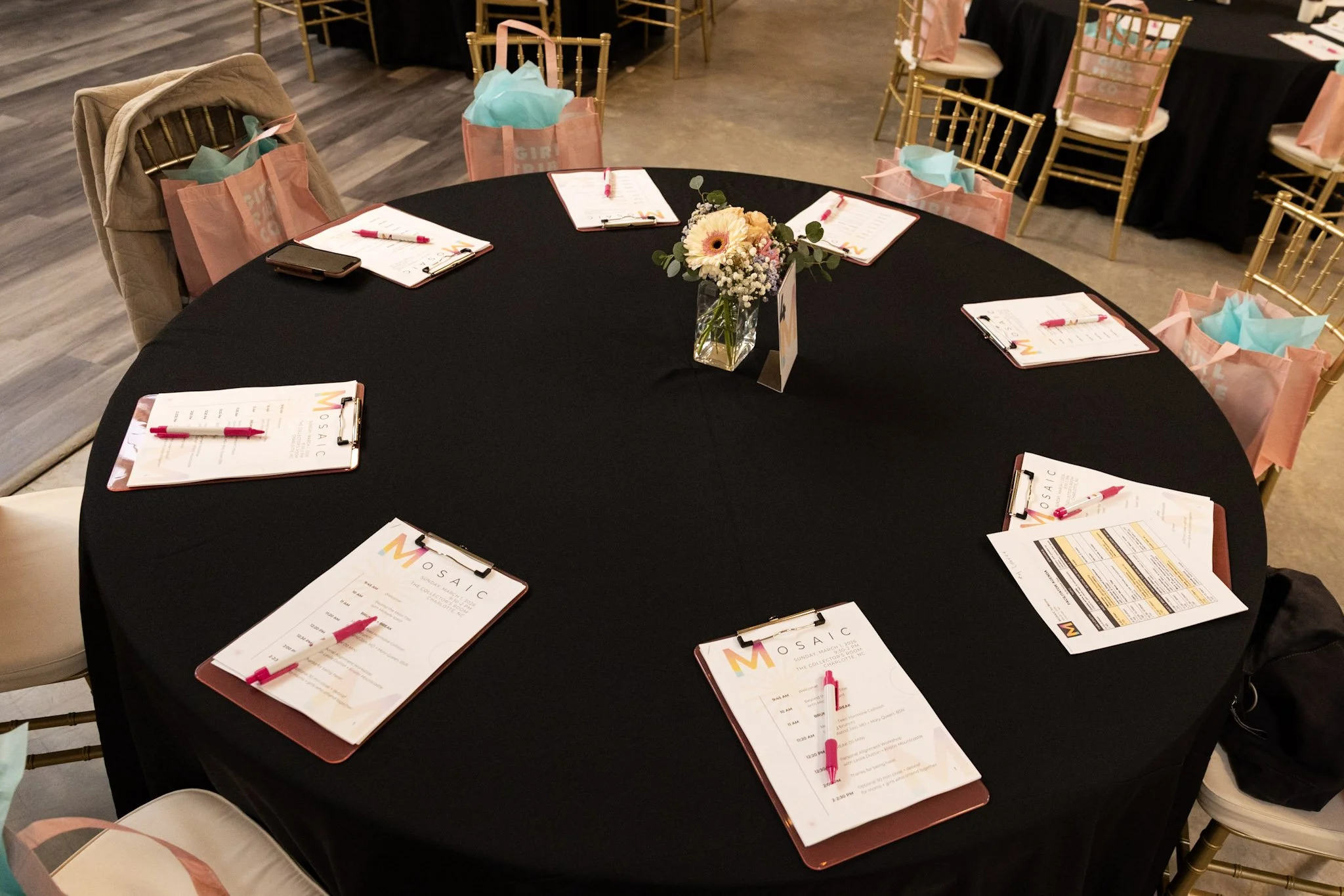 Round table set for a event with black tablecloth, pink and blue gift bags hanging from gold chairs, and clipboards with menus and pink pens. A small floral arrangement in the center.