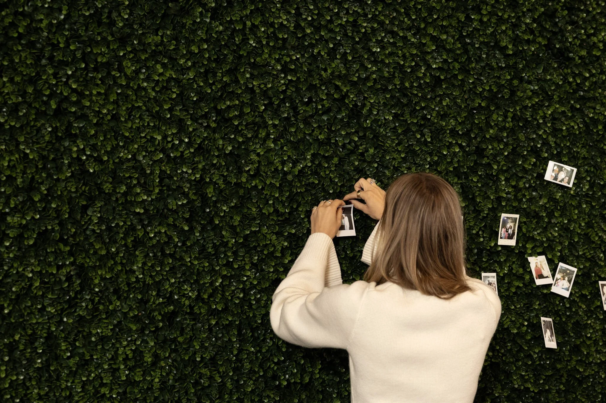 A woman with light brown hair wearing a beige sweater, taking a photo of her friend, against a large green leafy wall with several Polaroid photos attached to it.