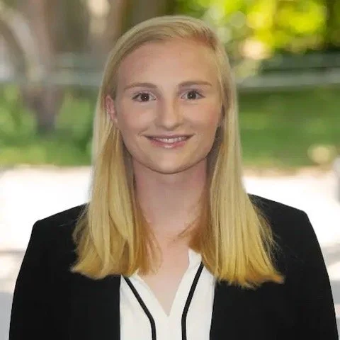 A young woman with blonde hair, smiling, wearing a black blazer over a white shirt, standing outdoors with blurred greenery in the background.