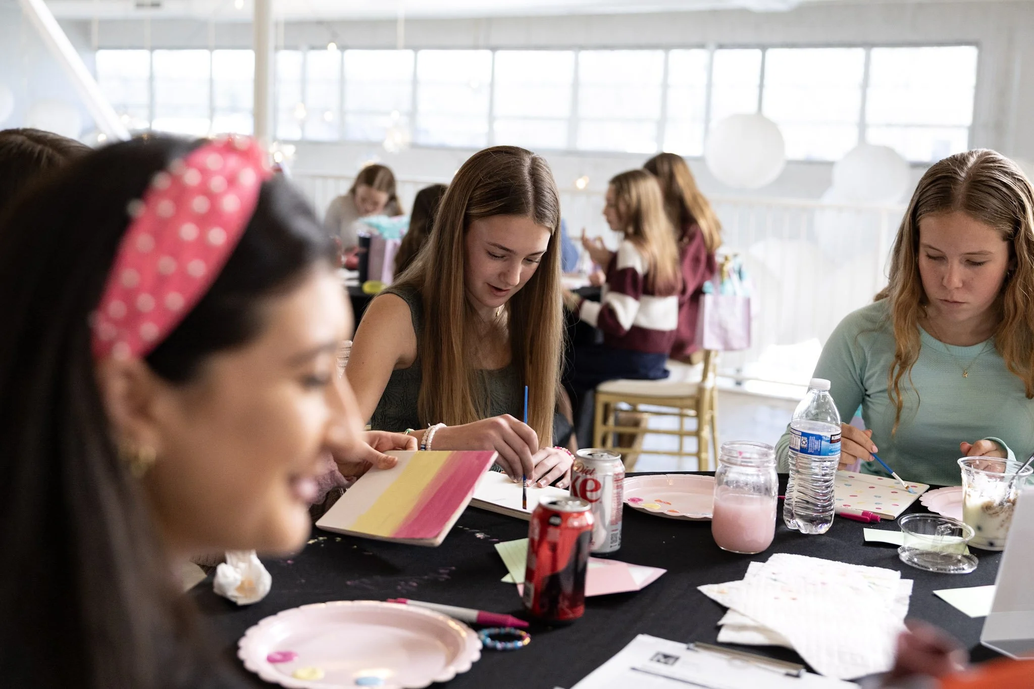 Young women sitting around a table at a birthday party, with gift bags, soda cans, water bottles, and plates in front of them, in a bright room with large windows.