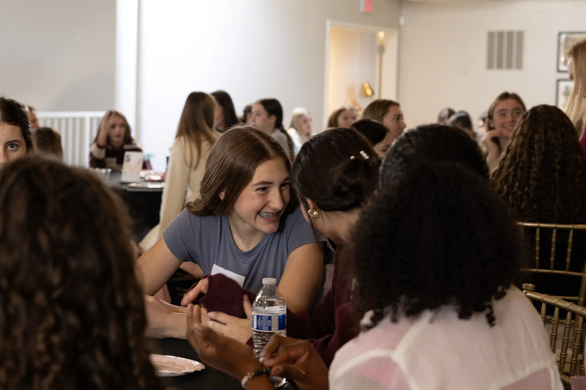 Young woman with short brown hair and braces smiling and talking to an older woman with glasses at a crowded indoor event.
