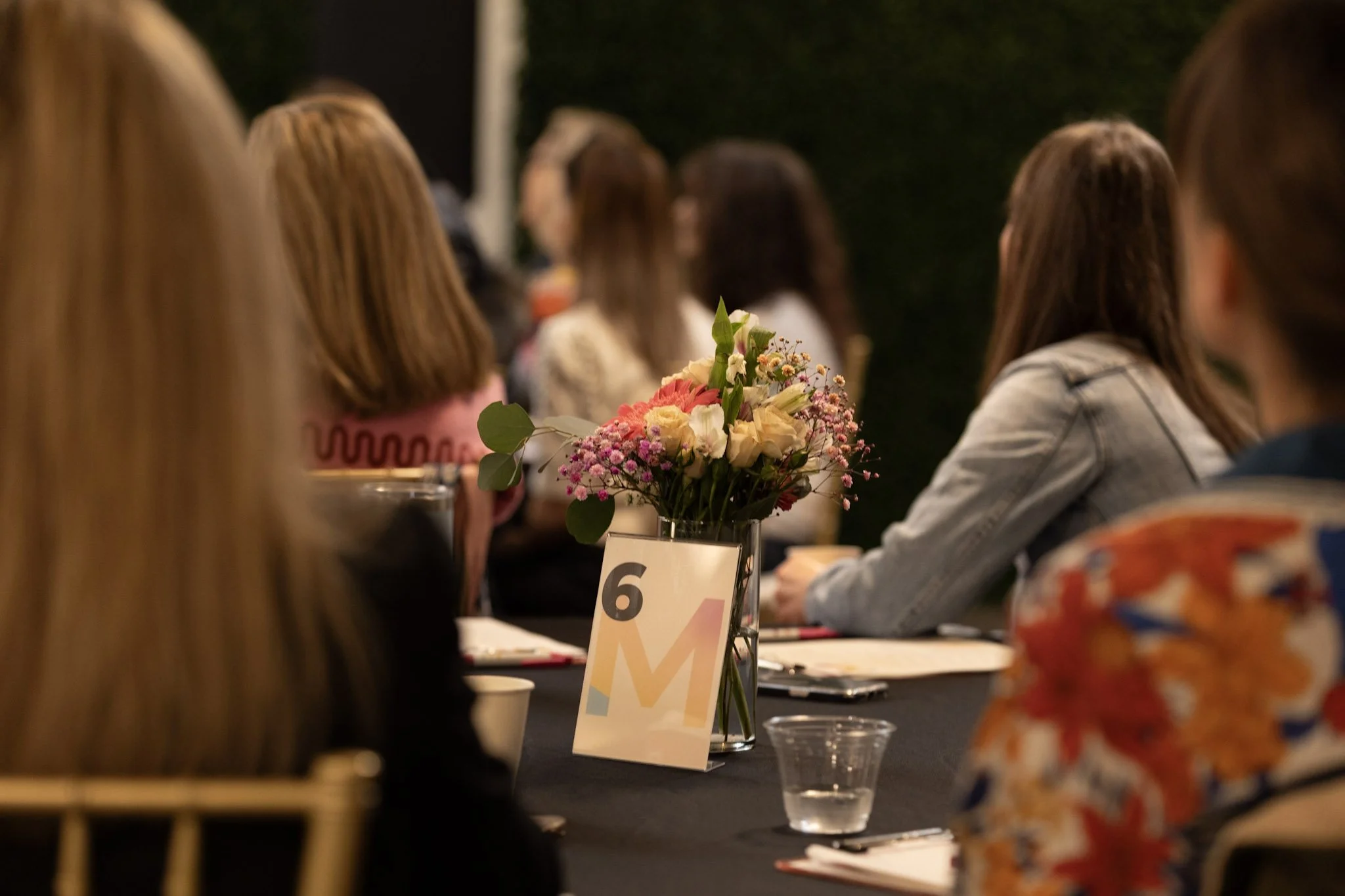 A blurred view of a group of people attending an event, with a focus on a flower arrangement on a table labeled '6' in the foreground.