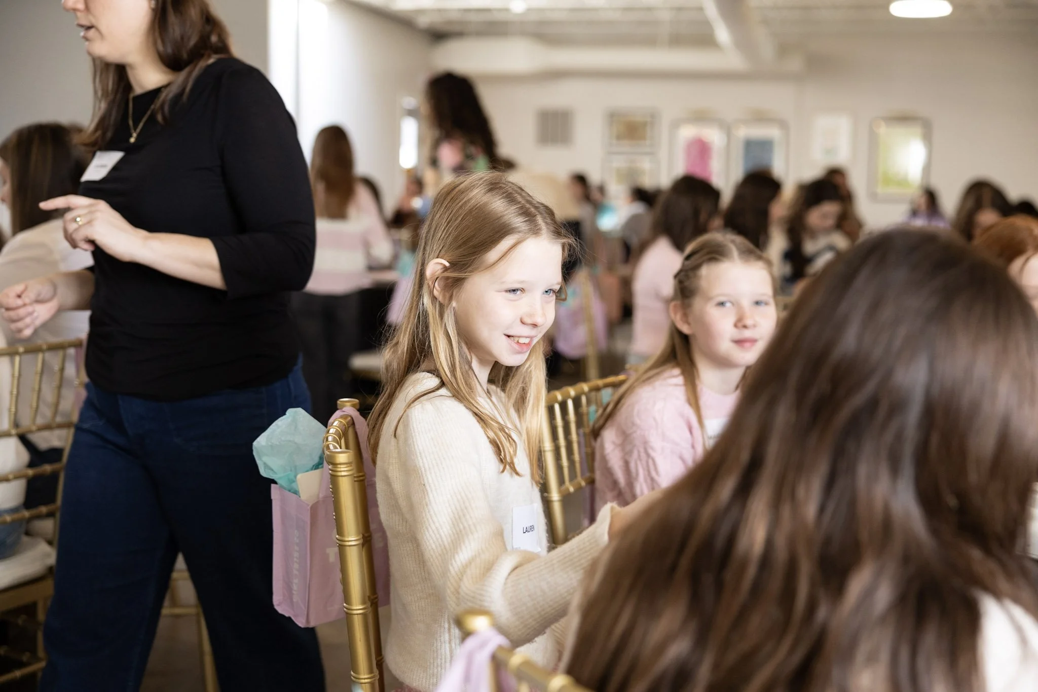 Group of young girls sitting at a long table in a well-lit room, possibly during a workshop or event, with some women standing and walking around.