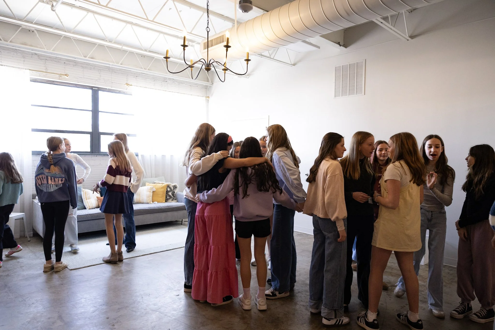 A group of young girls are gathered together inside a bright, spacious room, engaging in a social activity or game. The room has a modern, minimalist design with white walls, large windows allowing natural light, and a decorative chandelier hanging f