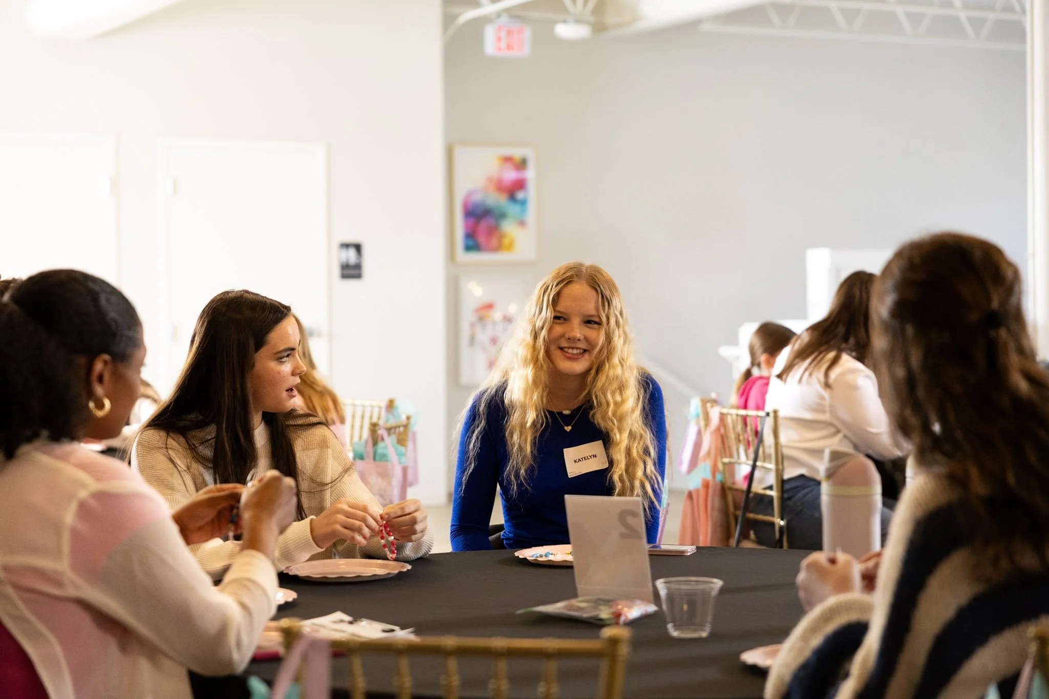 Group of women sitting around a table chatting at a social event or workshop, decorated with pink gift bags and tableware.