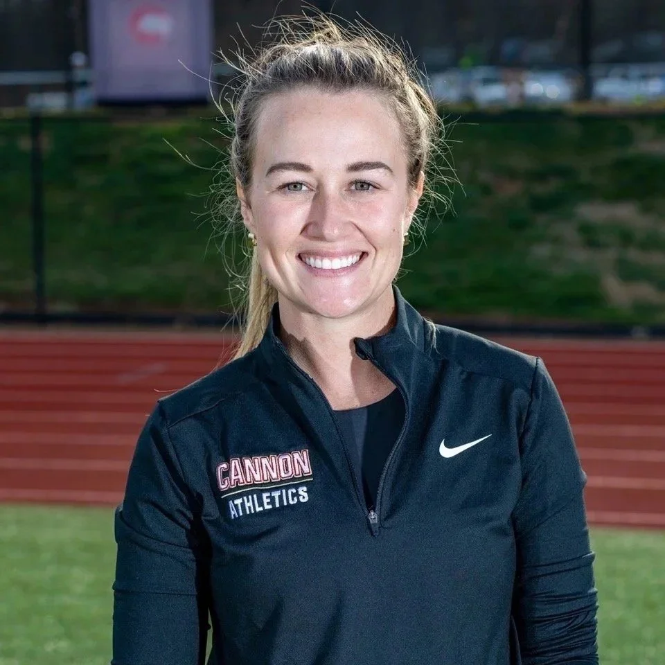 A smiling woman in a navy blue athletic jacket with a Nike logo, standing on a running track at sunset.
