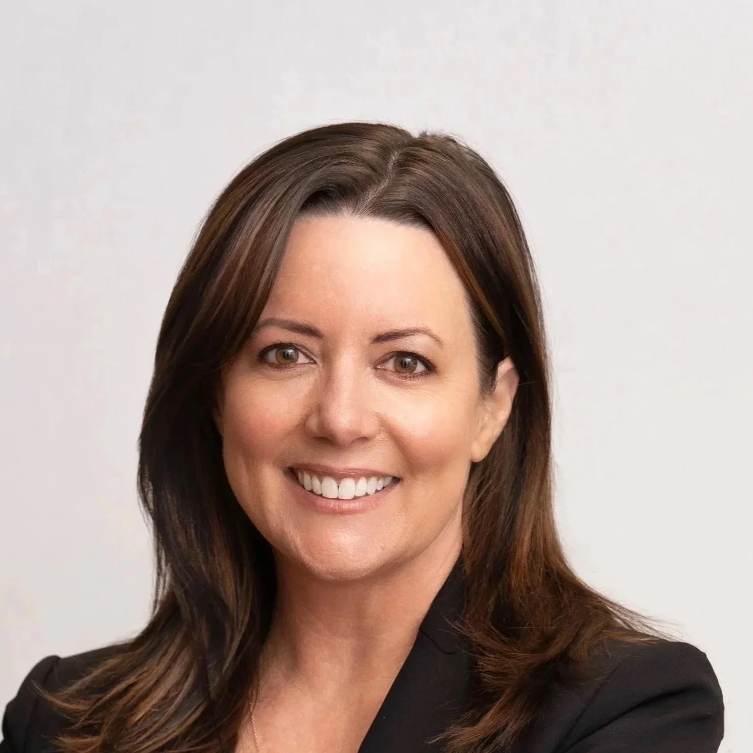 A woman with long brown hair, wearing a black blazer, smiling, against a plain white background.