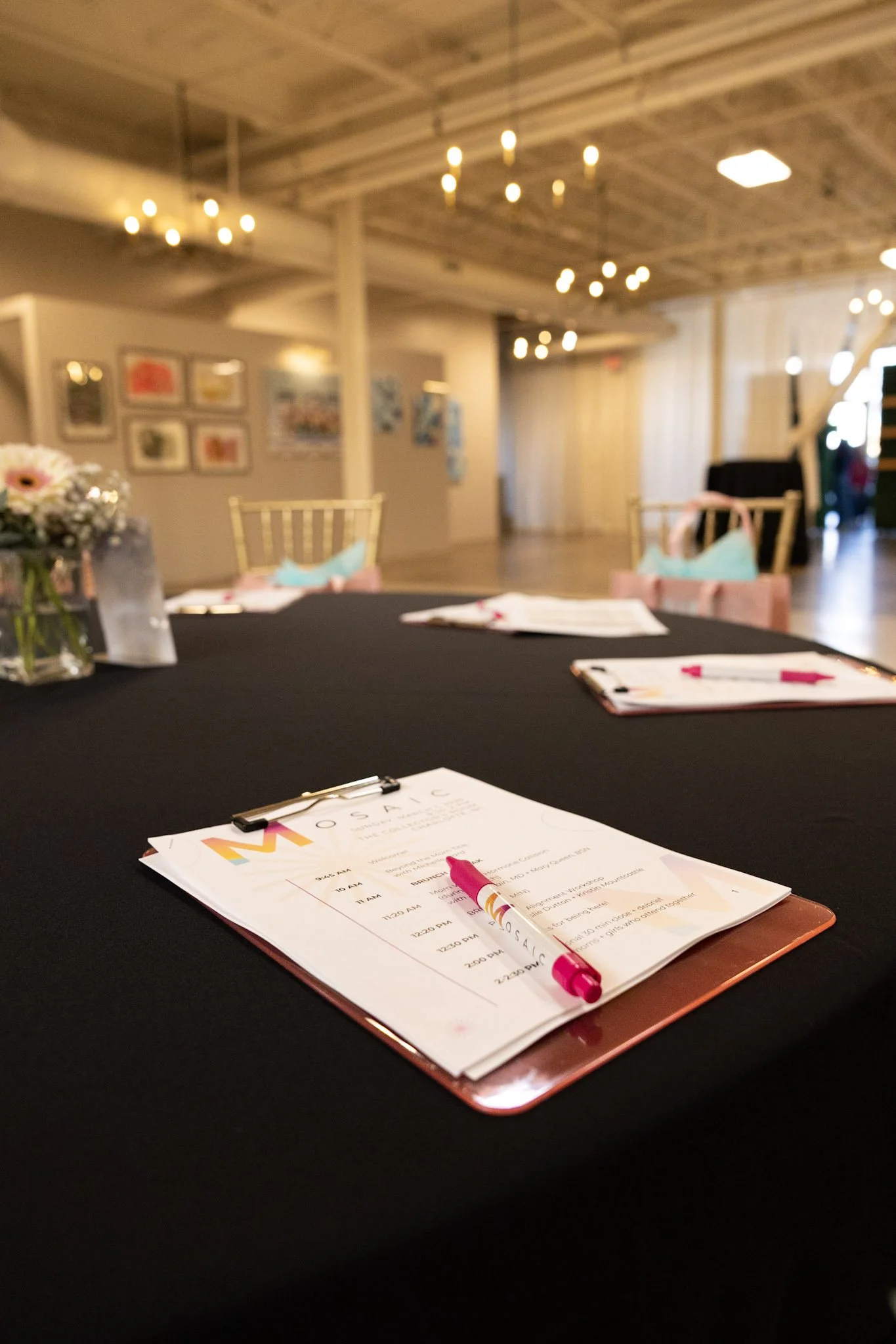 A conference or meeting room with a black table, flower arrangement, and printed agendas or programs. The background shows a wall with framed artwork and warm lighting, wood-paneled ceiling, and chairs with light-colored upholstery.