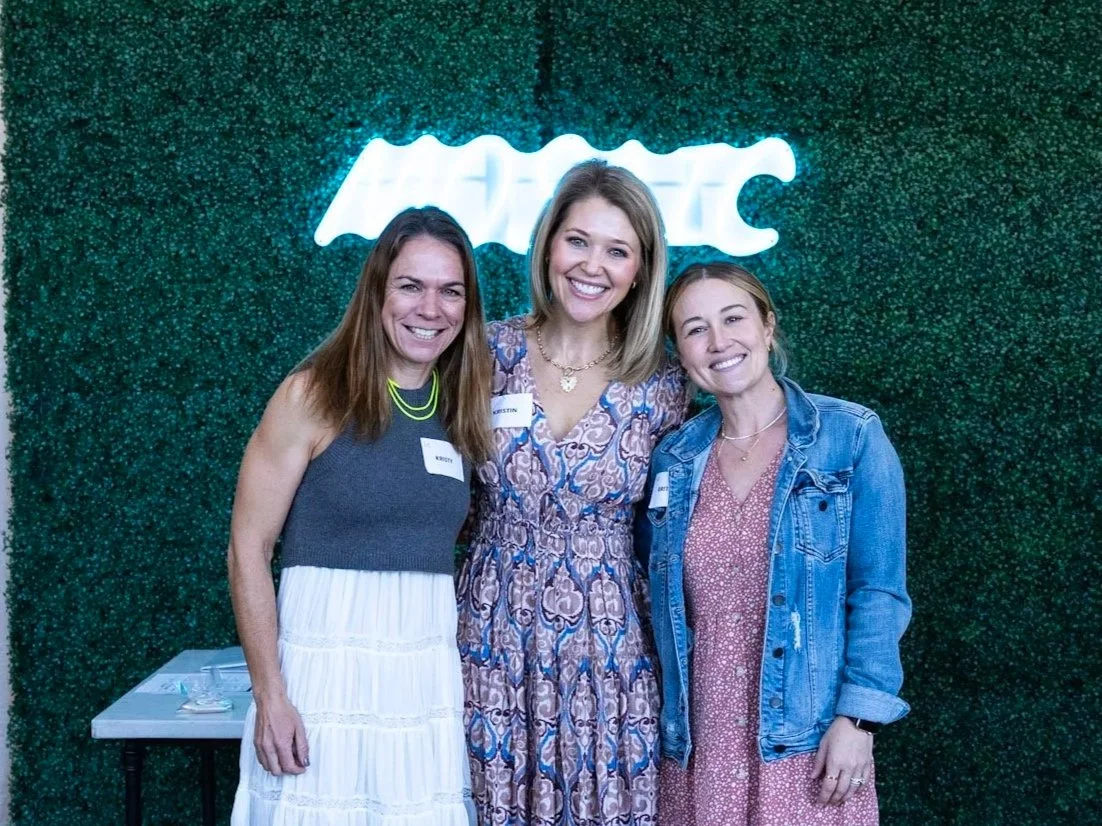 Three women smiling and posing together in front of a green wall with a neon sign. One woman is wearing a sleeveless top and a white skirt, the middle woman is in a patterned dress, and the woman on the right is in a pink dress with a denim jacket.
