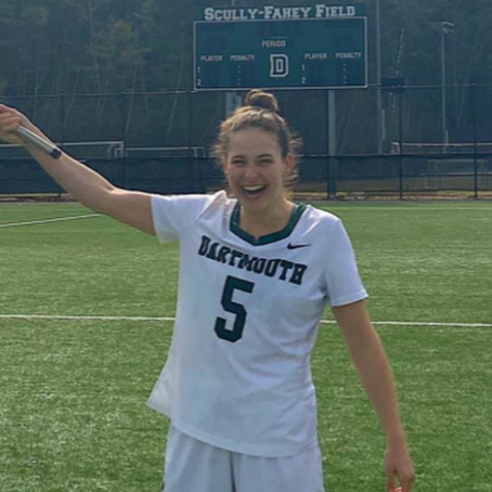 Young female soccer player smiling on a field, wearing a white Dartmouth jersey with the number 5, holding a hockey stick
