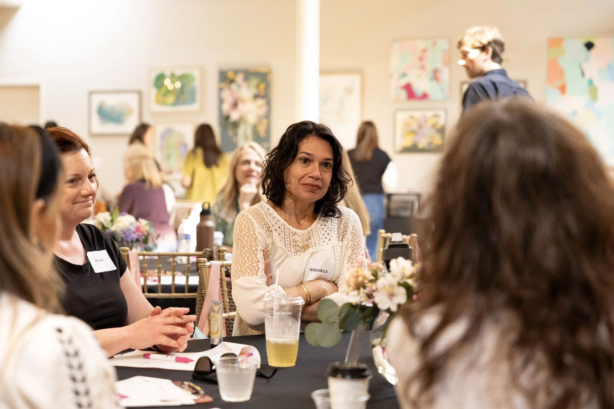 People sitting at a decorated table in an art gallery or museum, engaging in conversation during an event, with colorful artwork hanging on the walls.