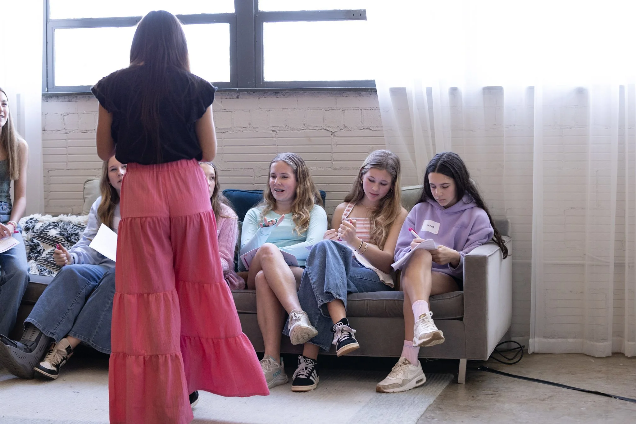 A girl with long dark hair wearing a black top and a pink skirt standing in front of a group of six girls sitting on a couch, engaging in a discussion. The room has large windows and white brick walls.