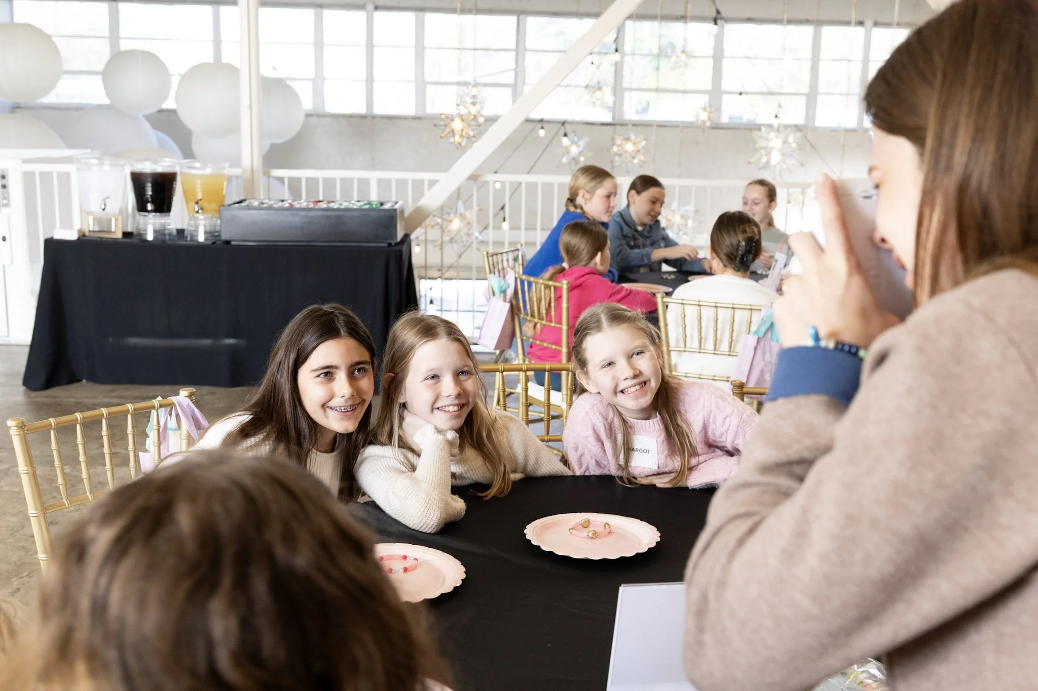 Group of children and a woman at a party, seated around a table with pink plates, in a bright room with large windows and hanging decorative lights.