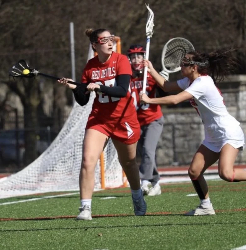 Two female lacrosse players in red and white uniforms are playing on a green field. One in red is holding a lacrosse stick with a yellow ball, while the other in white is reaching to grab the ball.